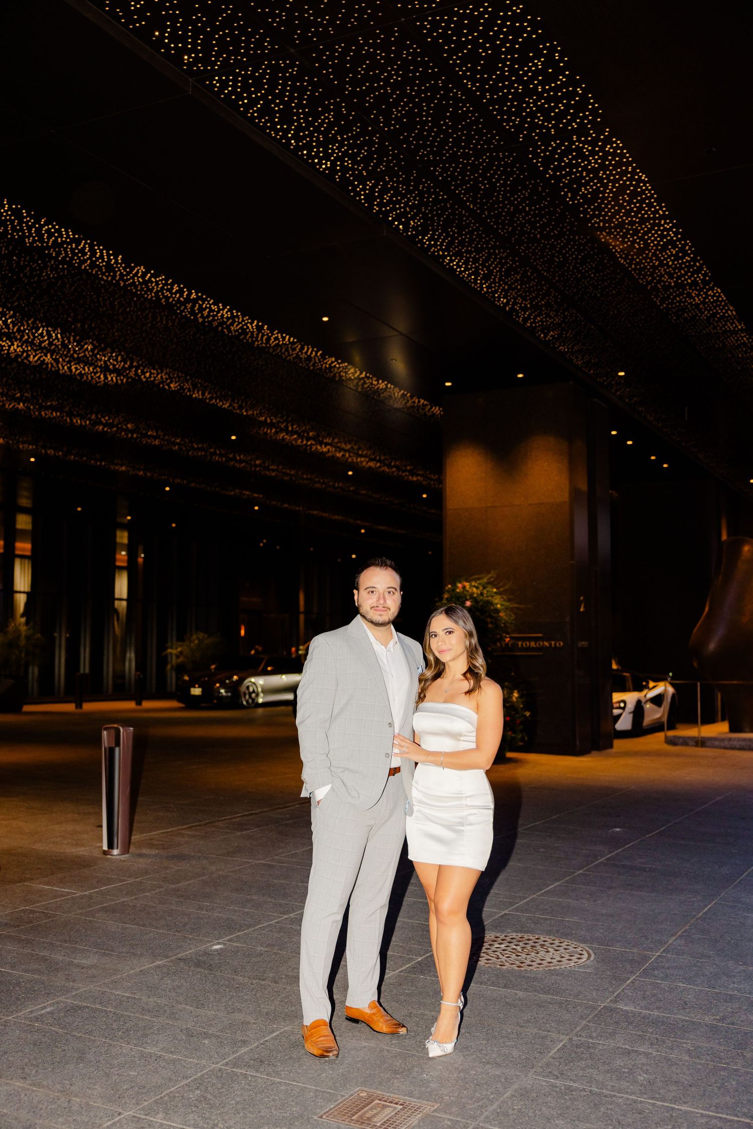 Couple poses under illuminated ceiling, with her in white dress and him in gray suit.