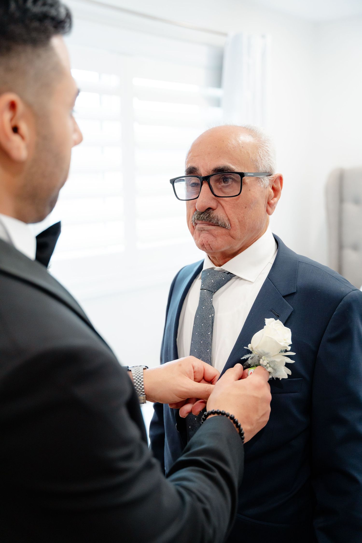 Two people in formal attire pin a white boutonniere flower on a navy suit jacket.