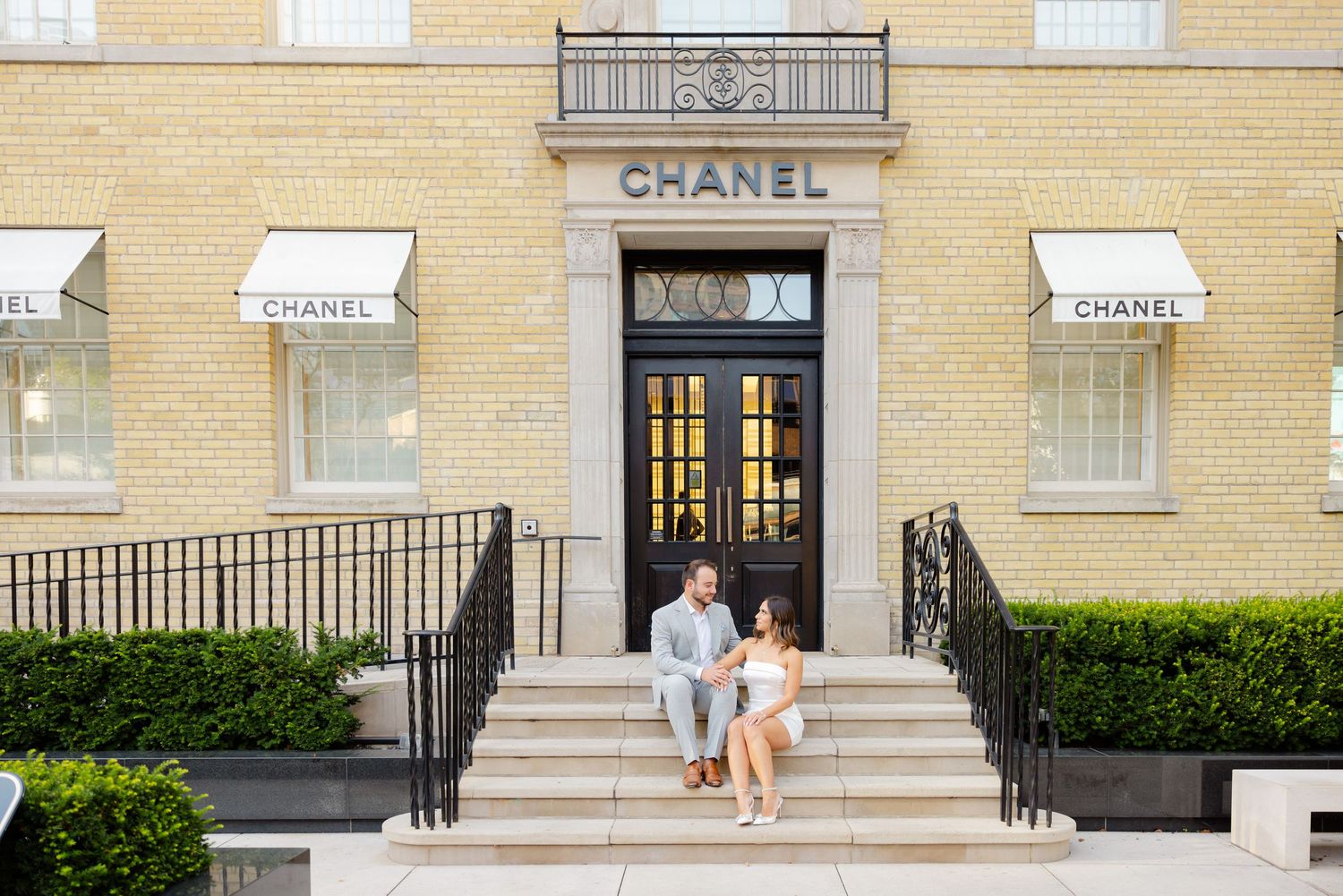 Wide view of couple on steps of Chanel store entrance with cream exterior and branded awnings.