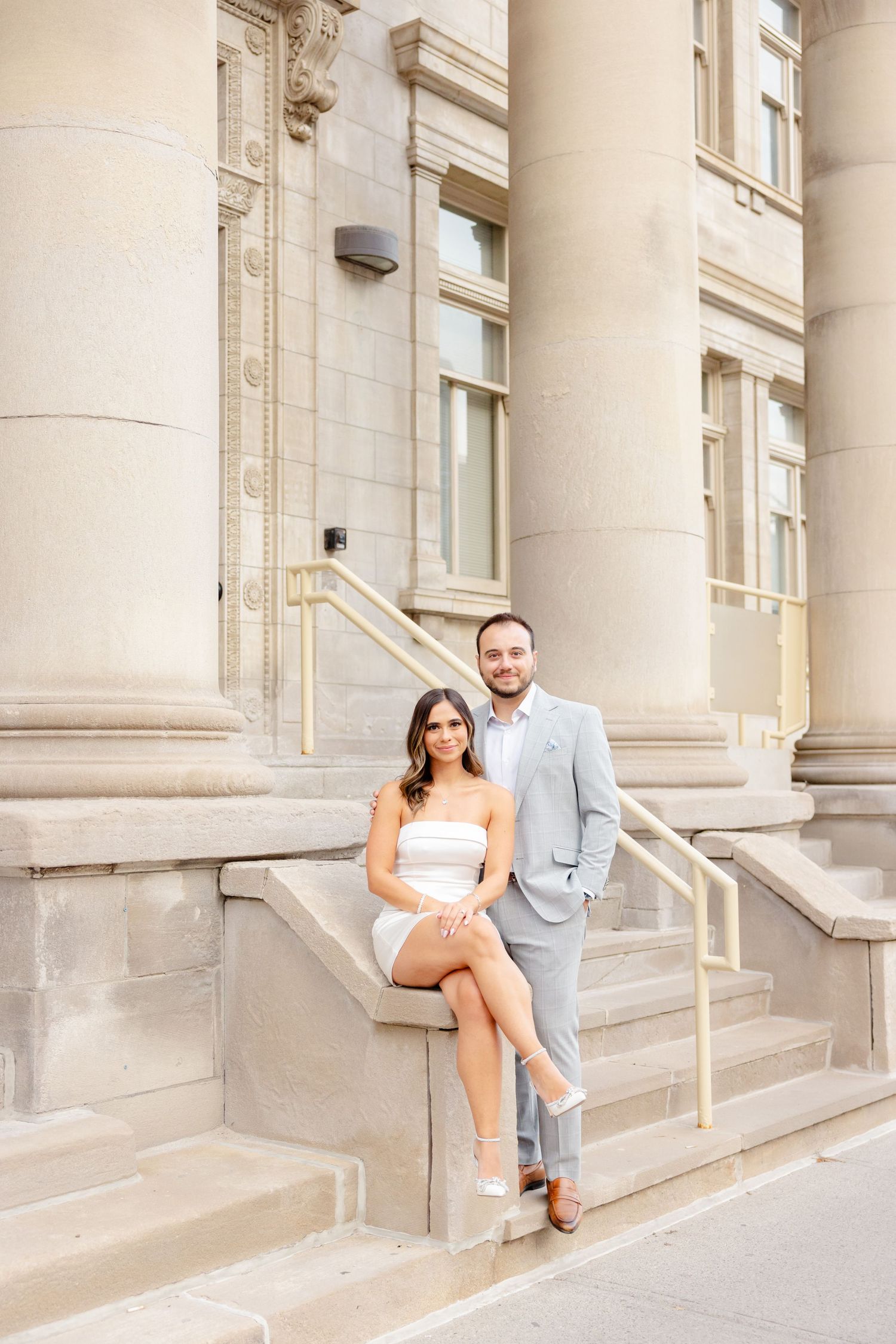 A couple poses casually on stone steps of a classic building with columns.