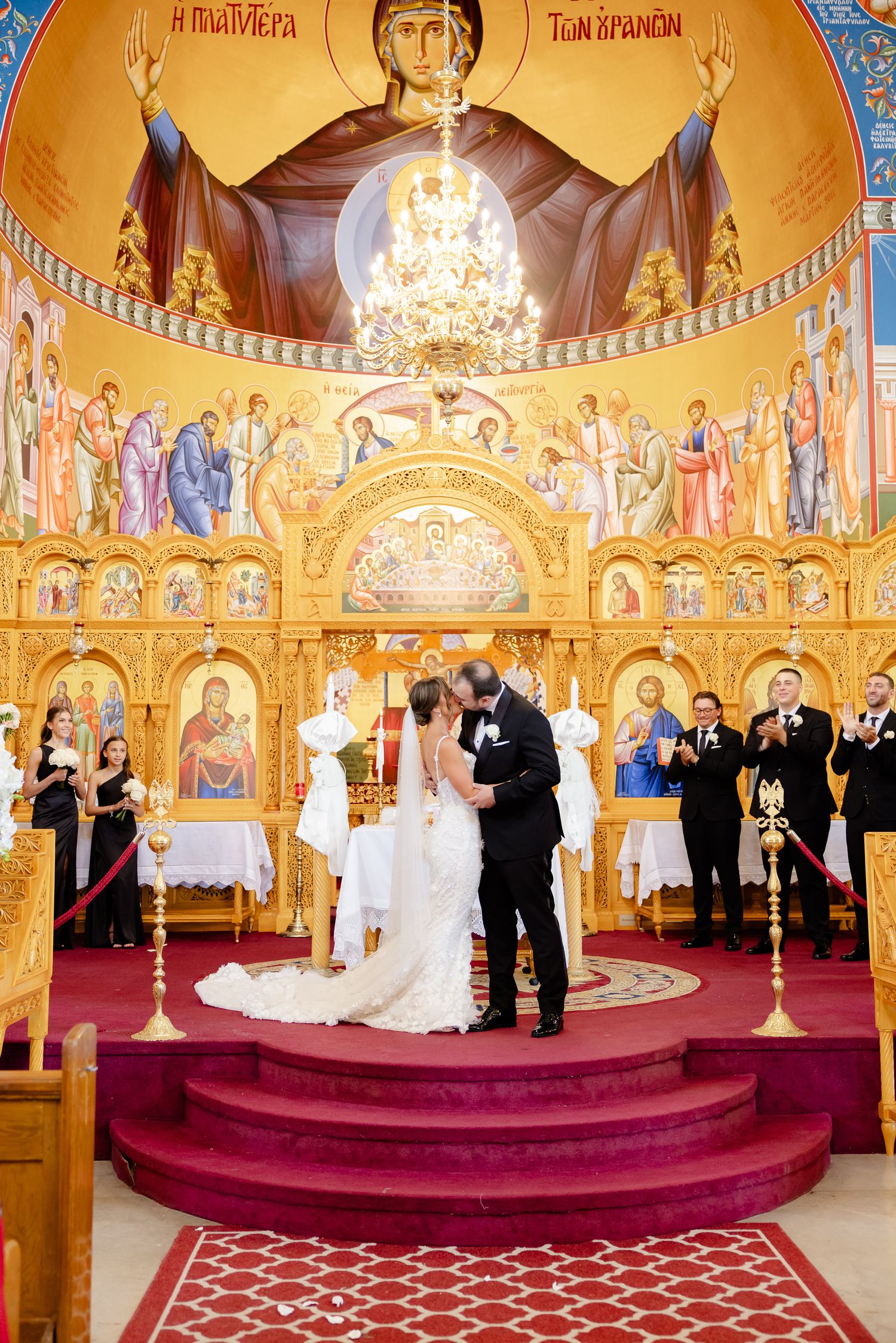 Orthodox church interior with golden walls and religious murals shows a wedding ceremony taking place at the altar.
