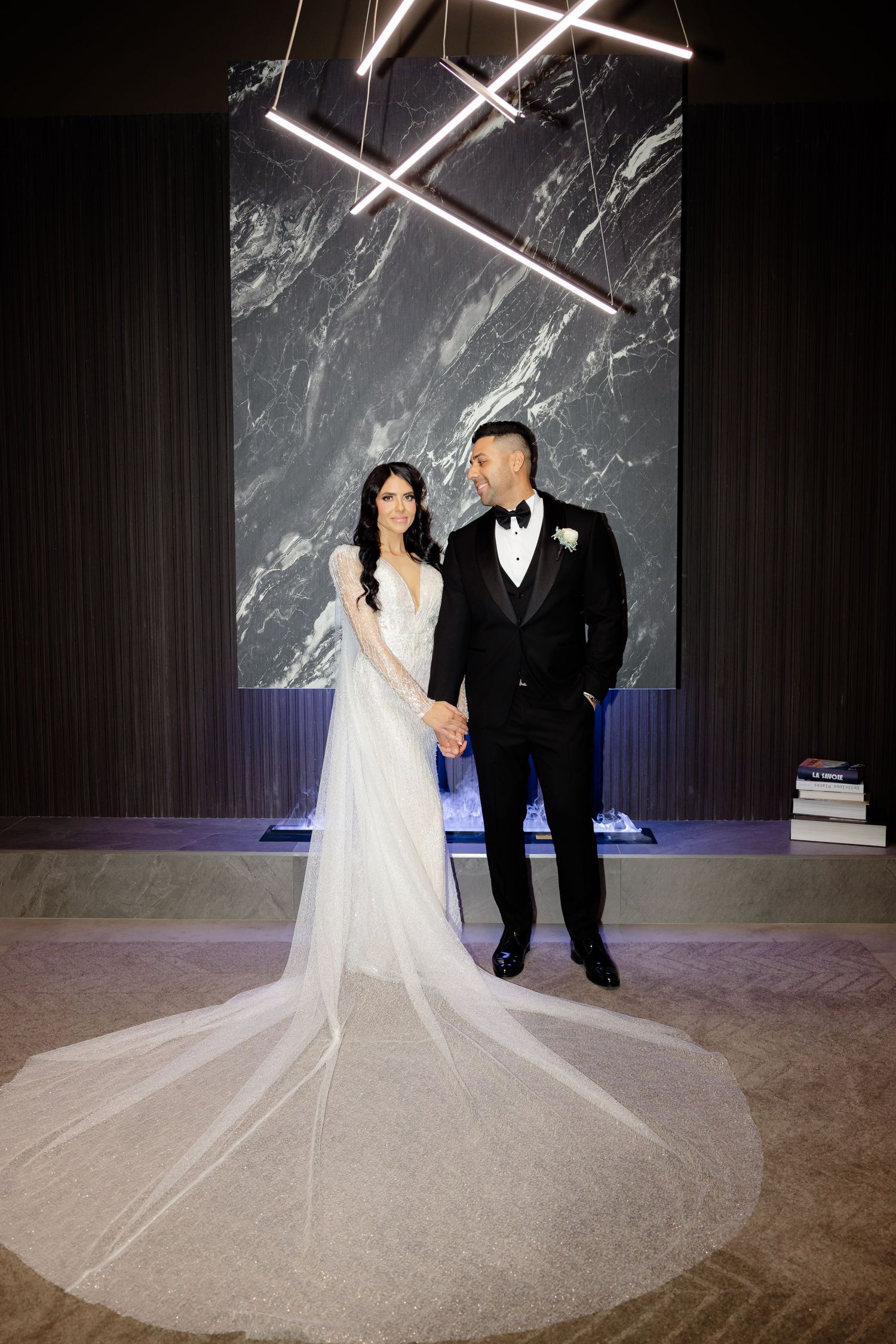 Elegant couple poses in formal wedding attire against modern marble backdrop with geometric lights.