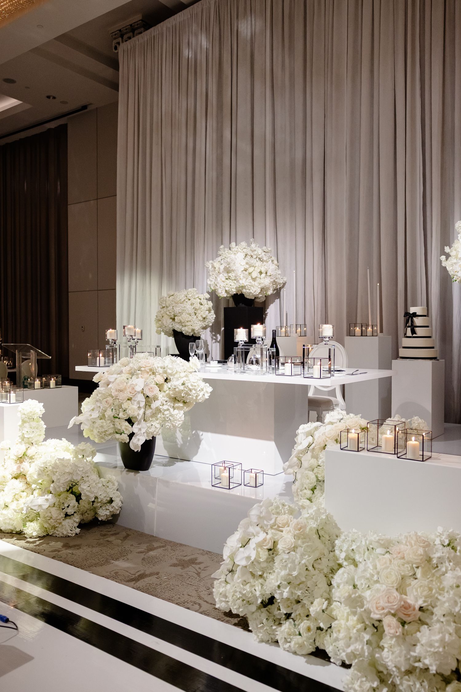 Elegant wedding decor display with white floral arrangements and tiered platforms against a floor-length curtain backdrop.
