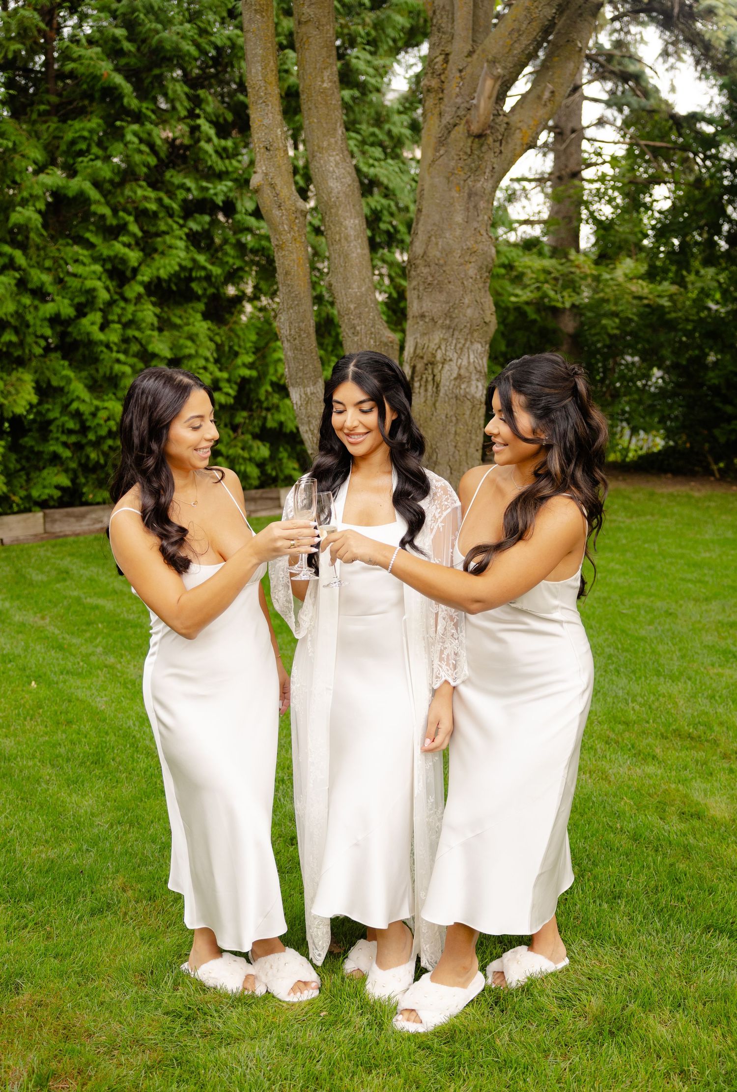 Three friends in white dresses laugh and celebrate together in a lush green garden setting.