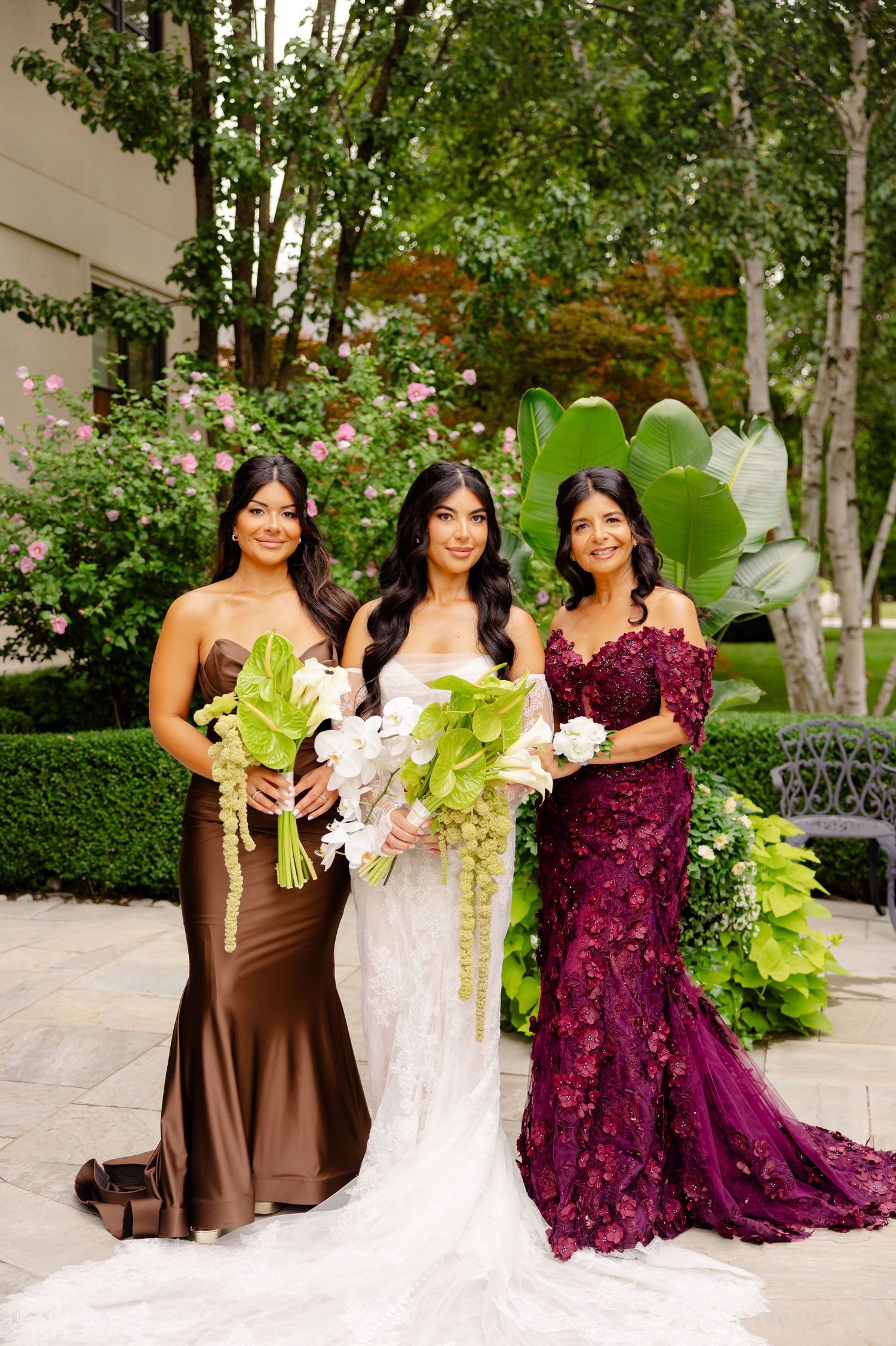 Wedding party poses in elegant brown and burgundy gowns alongside the bride in a garden setting.