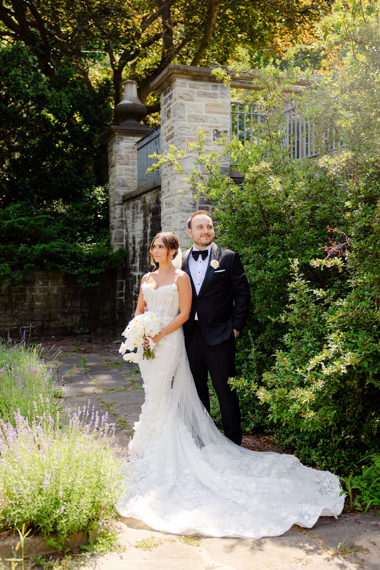 A couple in wedding attire stand together in a beautiful garden setting with stone walls and lush greenery.