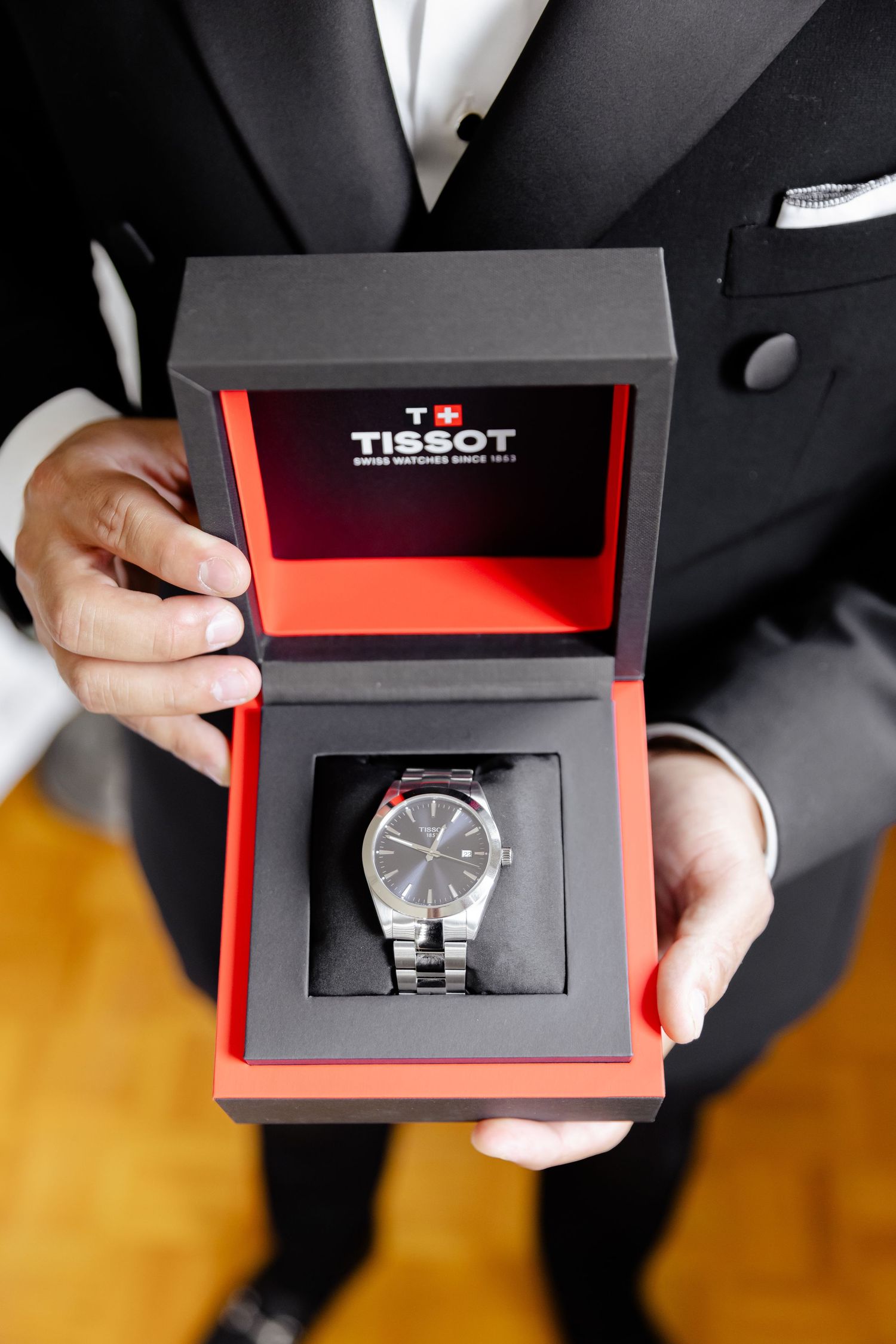 Sequence of hands holding and opening a Tissot watch presentation box against dark and yellow background.
