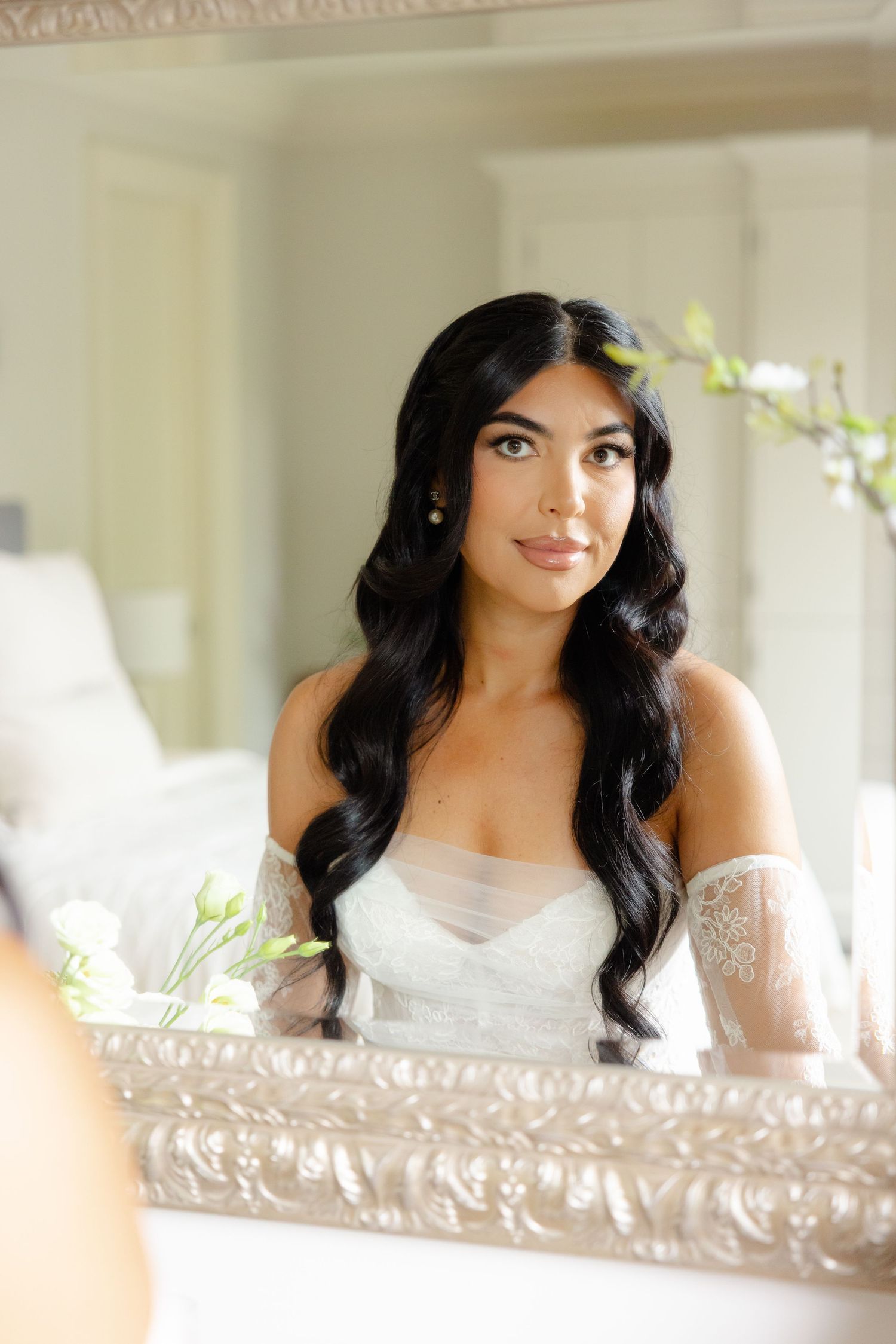 A woman in a white lace dress sits near a window with soft natural lighting for an elegant portrait session.