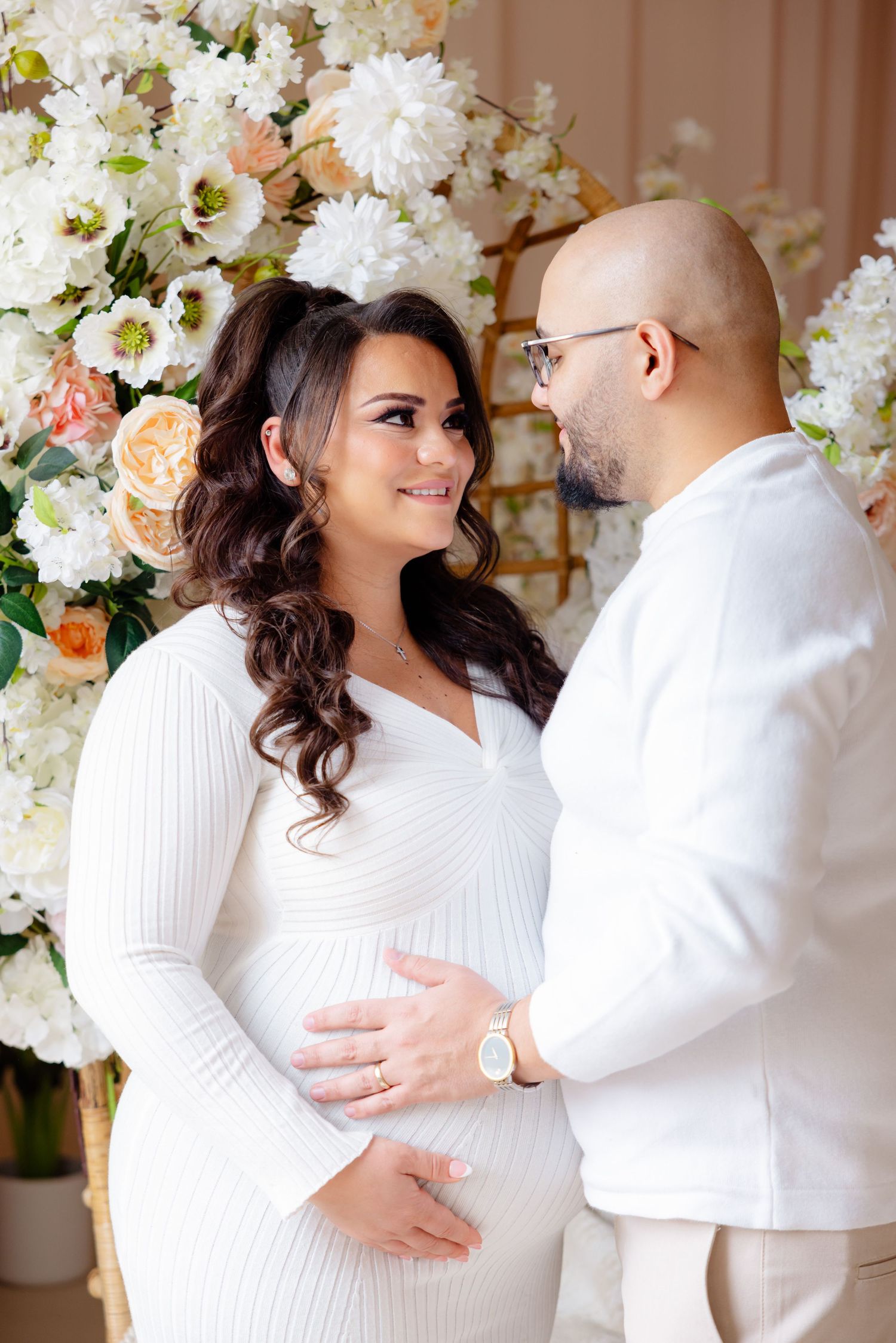 A couple in white outfits embraces in front of a floral backdrop of white and peach blooms during a maternity photoshoot.