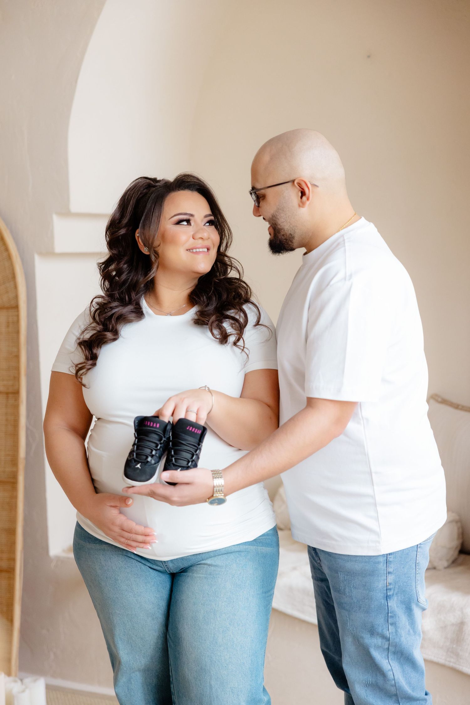 Couple in white shirts holding tiny baby shoes while standing together in a bright interior space.