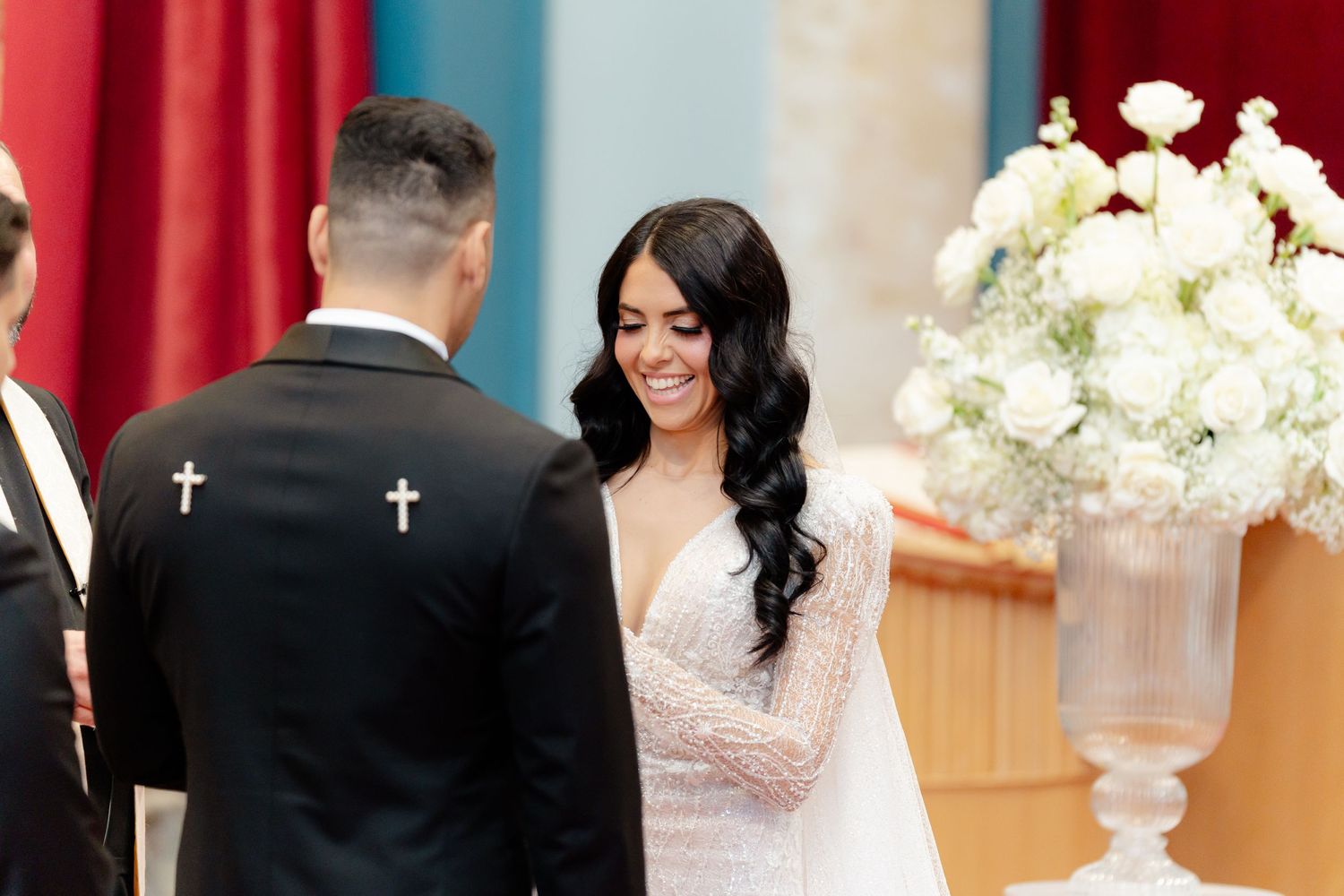 A bride in a white lace gown smiles at her groom during their wedding ceremony with floral decorations.