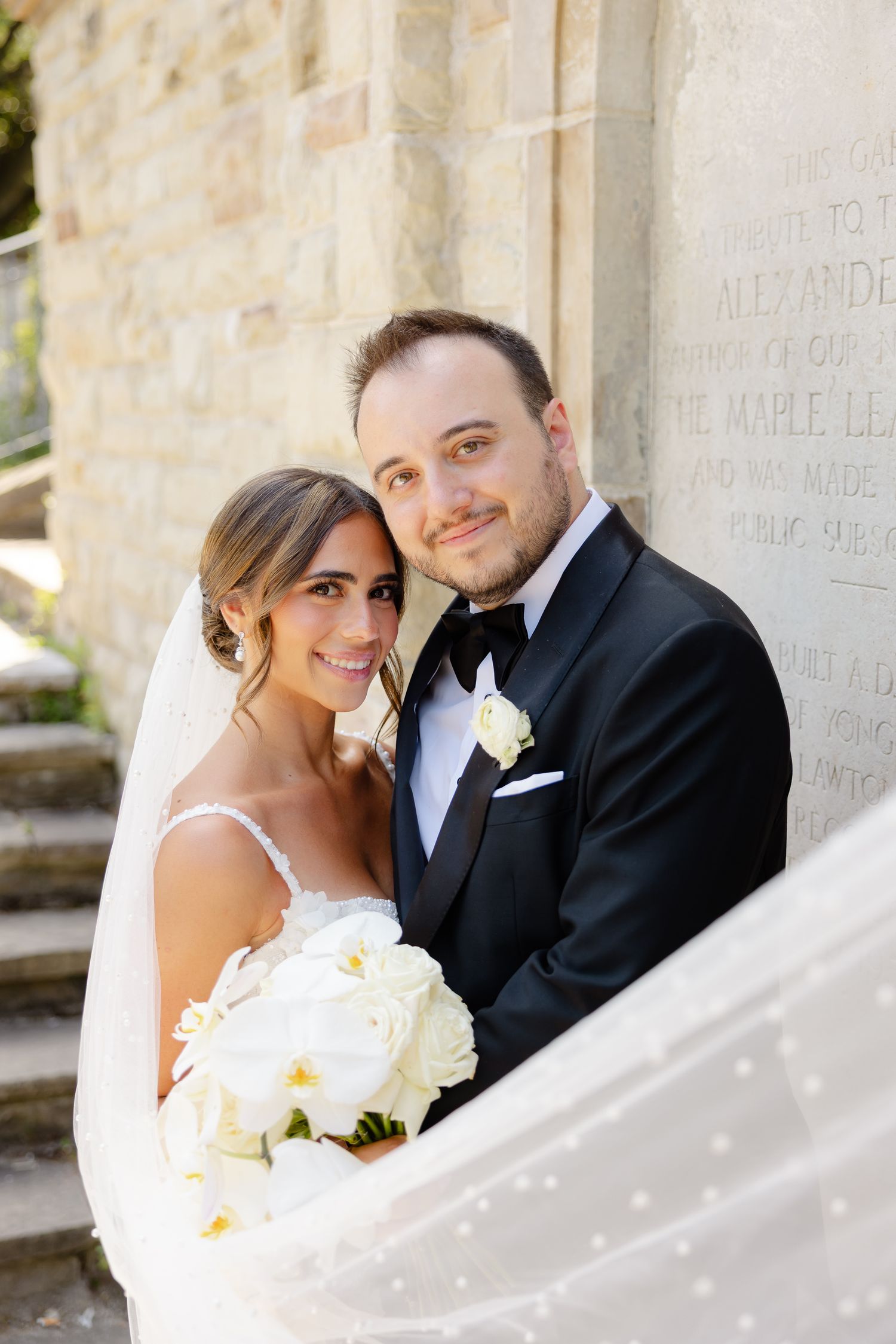 A bride and groom pose together against a stone wall, with the bride holding a white bouquet and wearing a traditional veil.