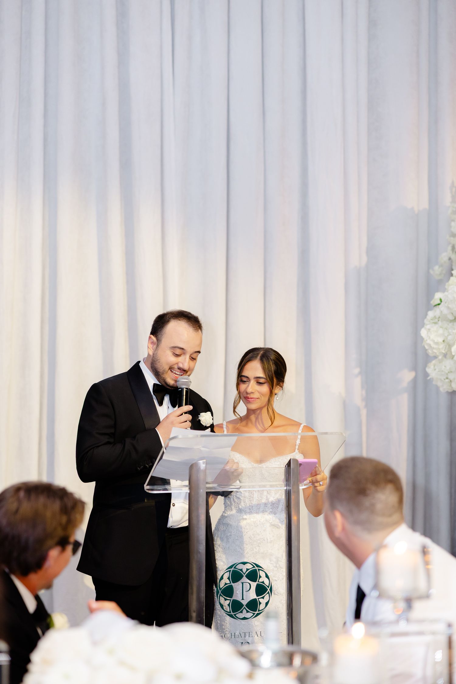 Two people stand at a podium giving speeches during a wedding reception with white curtains in the background.