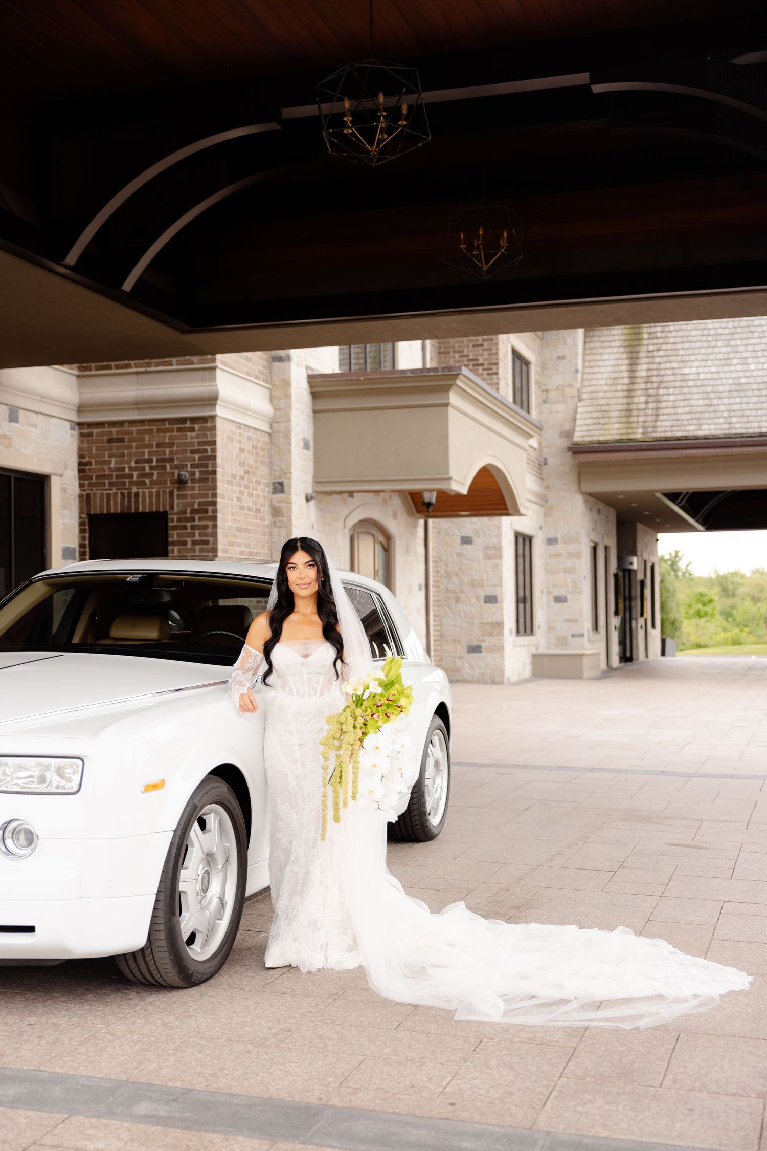 Elegant wedding couple posing with a white luxury sports car under a brick building portico.