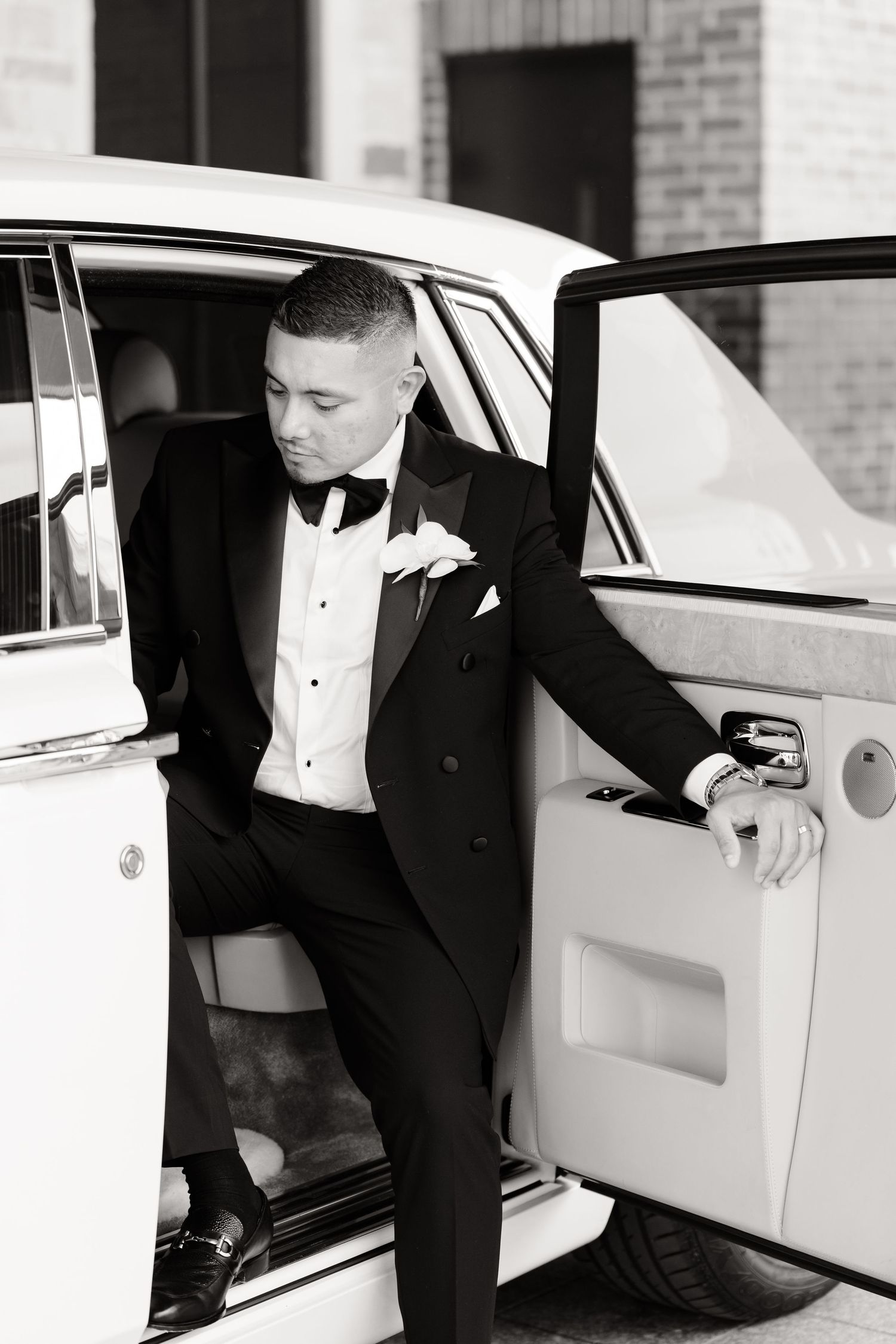 A groom in a classic black tuxedo enters a white luxury wedding car from multiple angles.