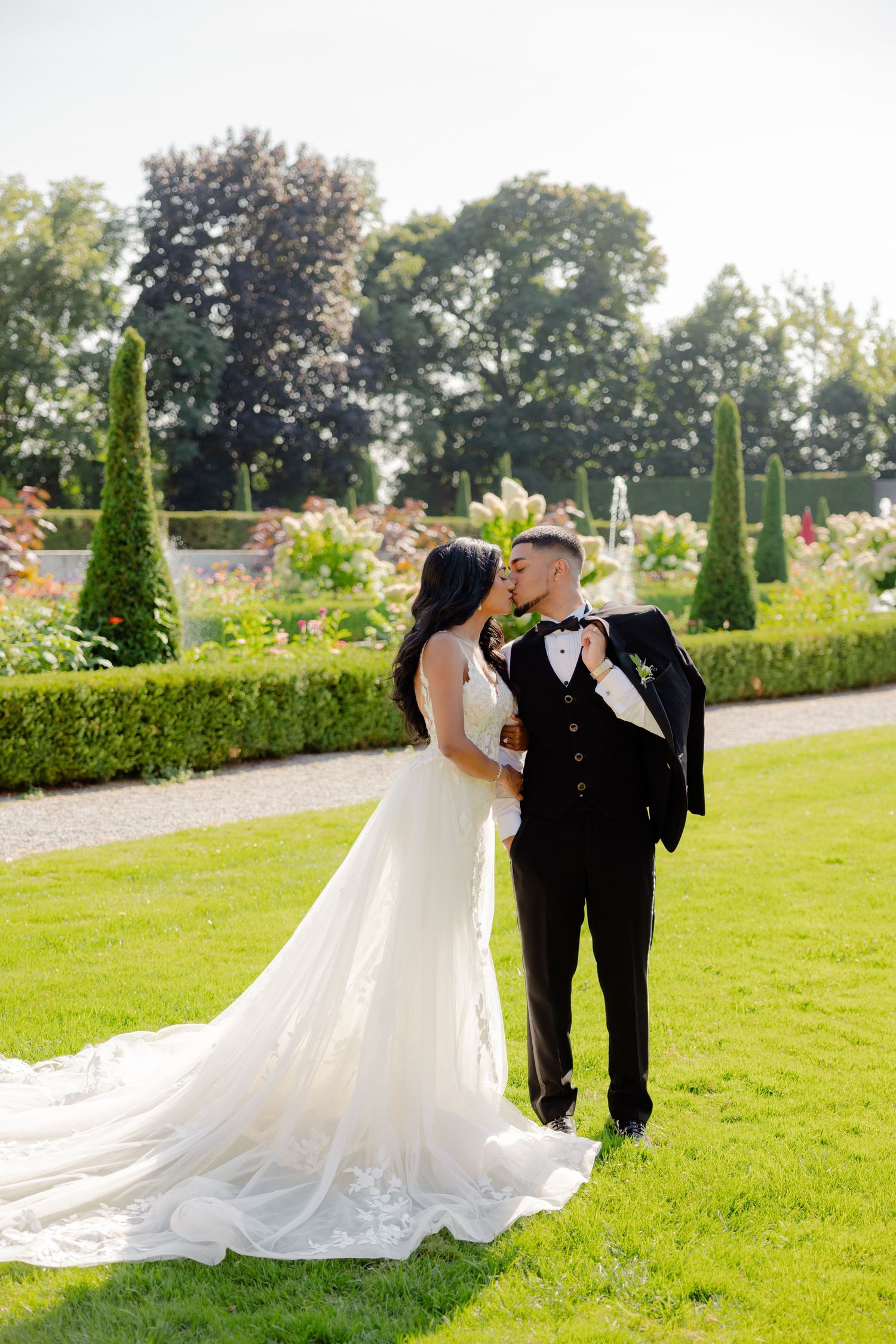 A romantic wedding photo in a manicured garden with tall topiary trees and colorful flowers in the background.