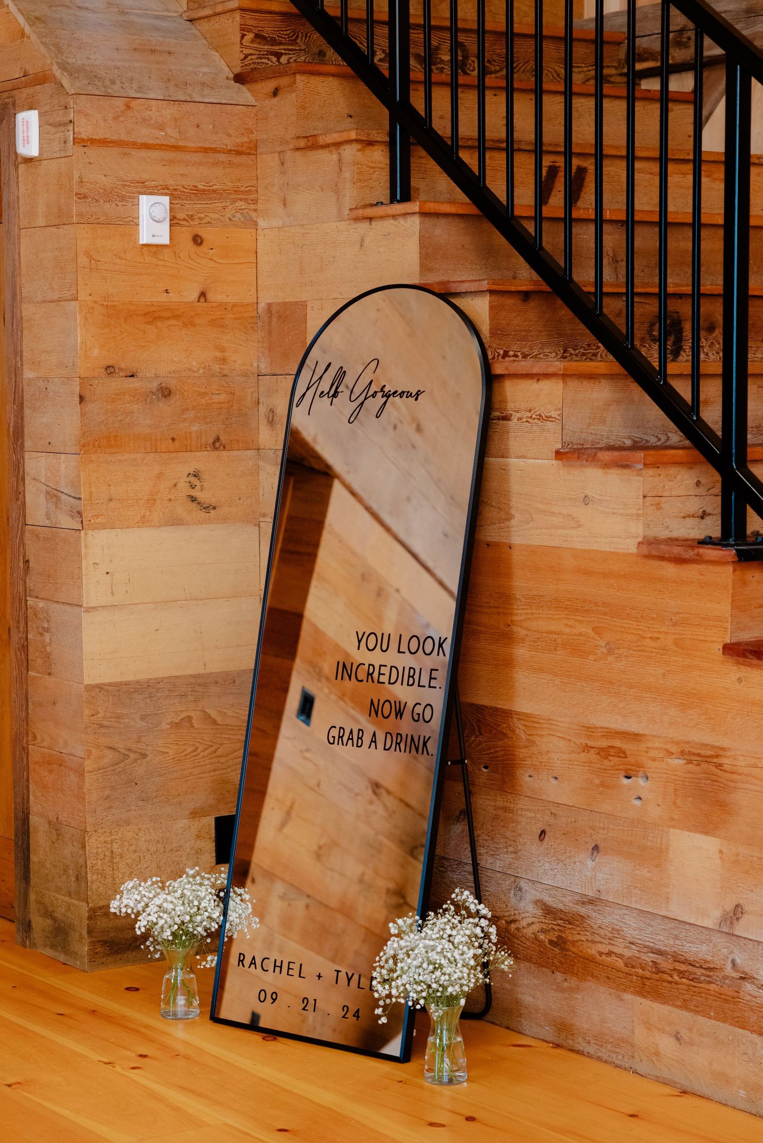 Mirror wedding sign with text leaning against rustic wooden wall with baby's breath flowers.