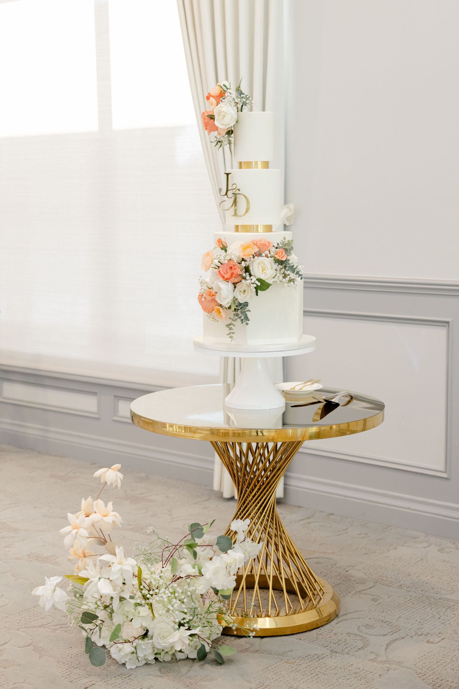 Elegant white wedding cake displayed on gold pedestal table with coral and white floral decorations.