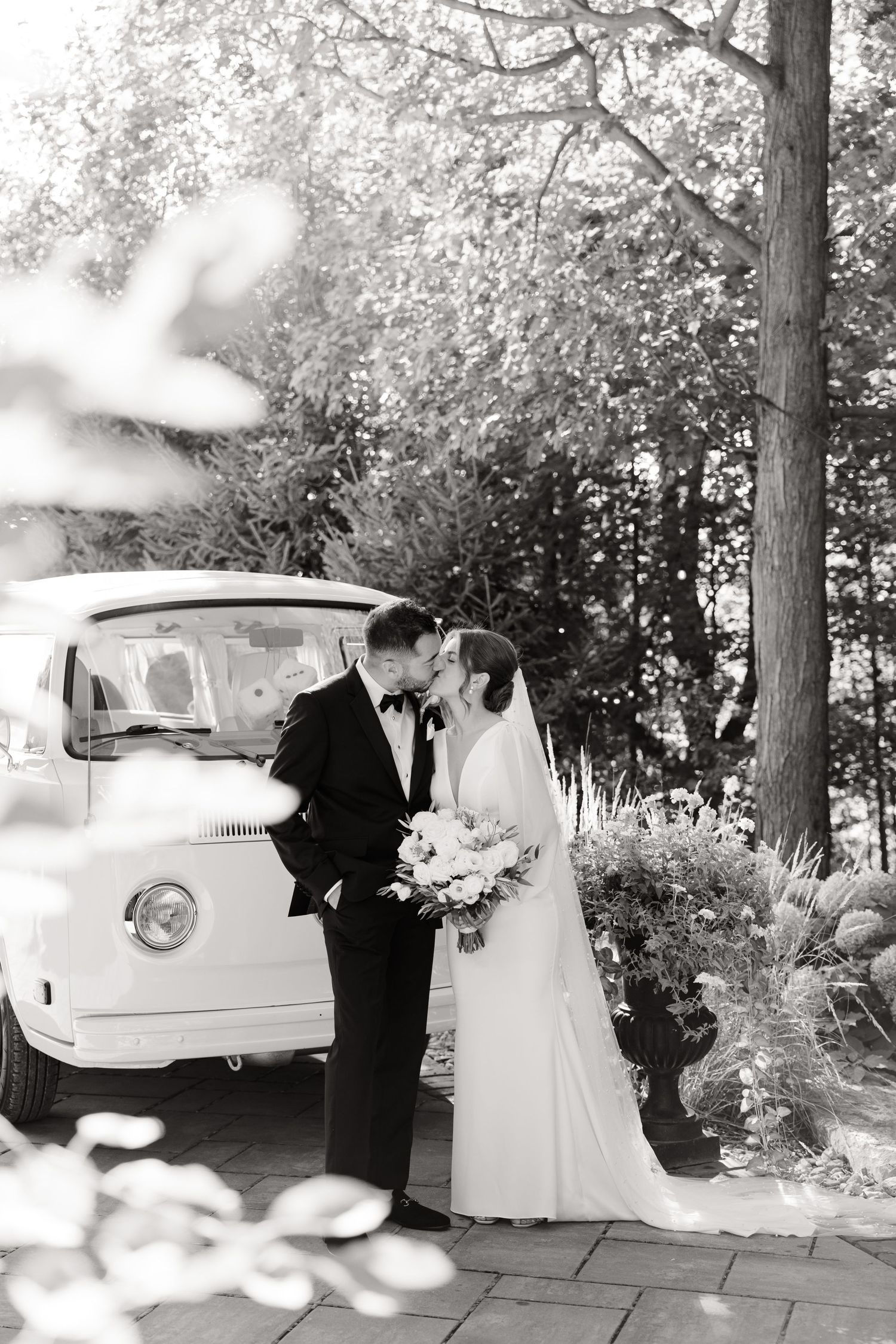 Black and white wedding photo showing a couple with a vintage van amongst trees and stone pathways.