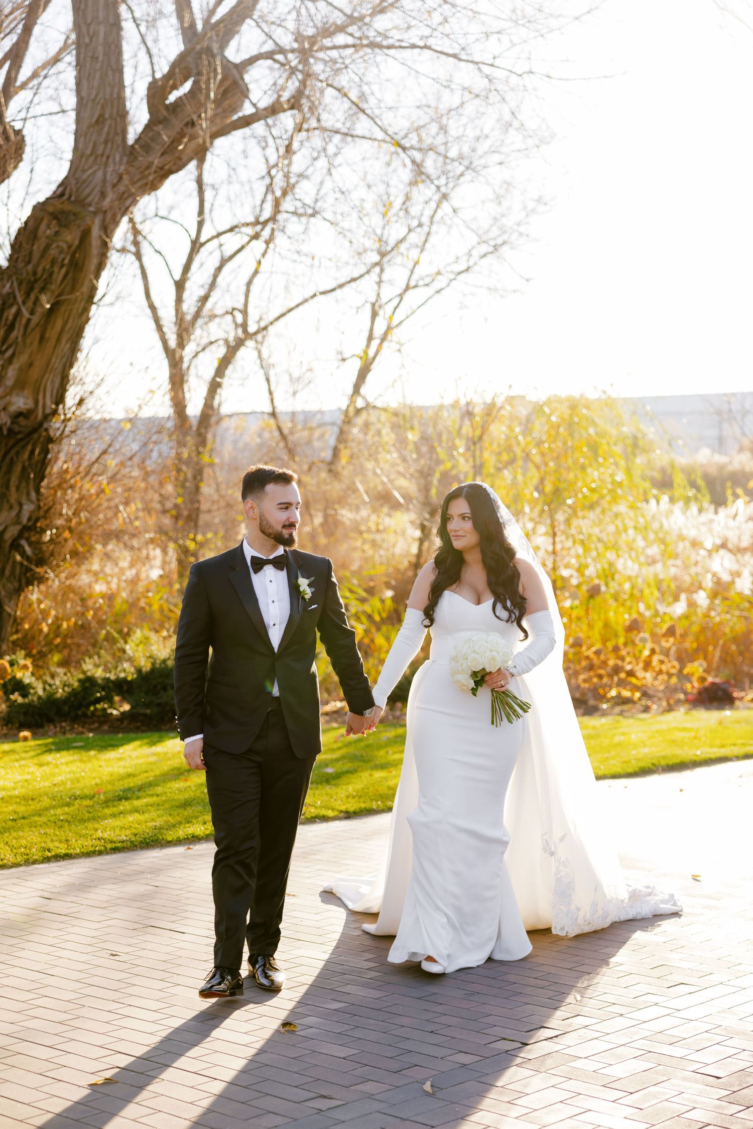 Wedding couple walking hand in hand on brick path with autumn foliage in background.