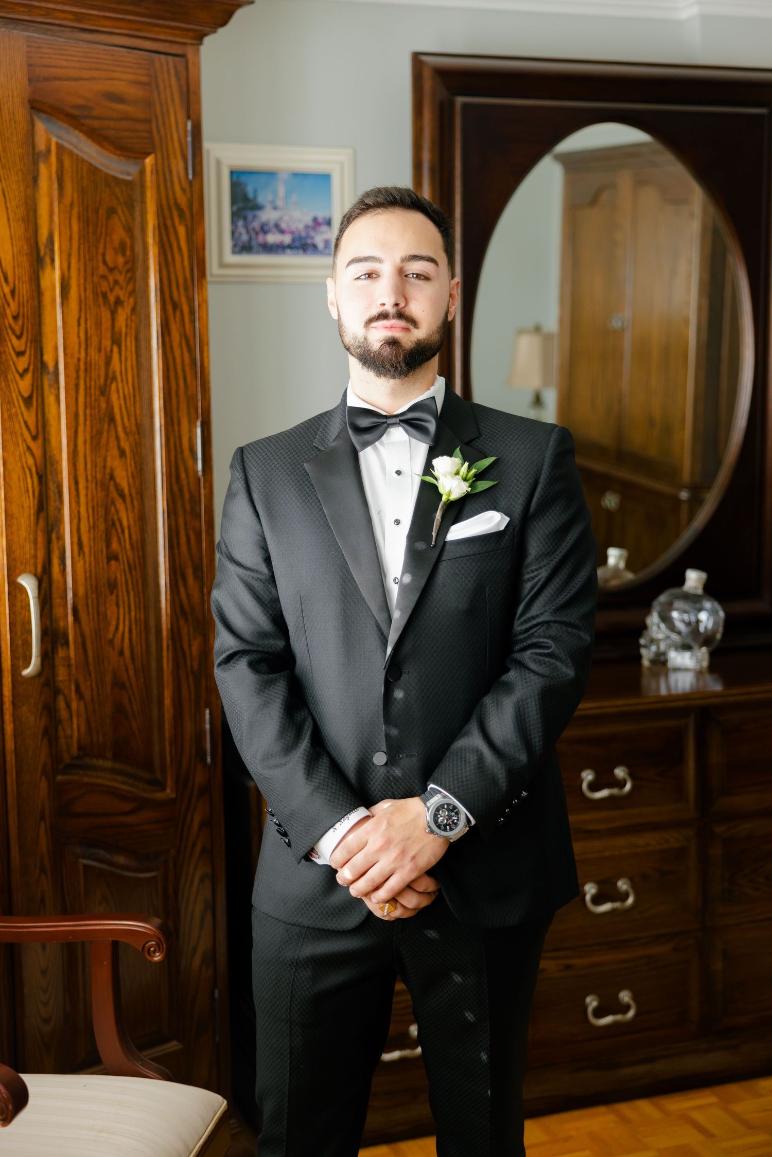 A groom in a classic black tuxedo with white boutonniere poses in front of an antique wooden dresser.