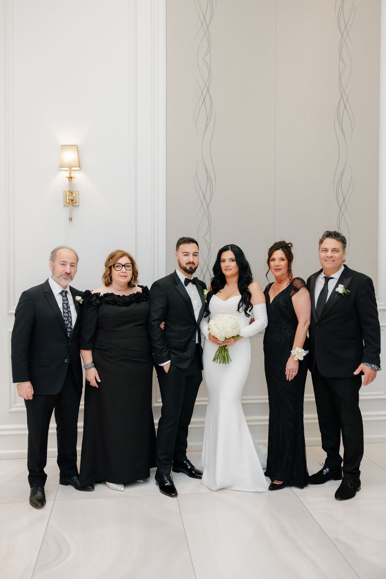 Extended family formal portrait at an elegant wedding venue with white walls and decorative lighting.