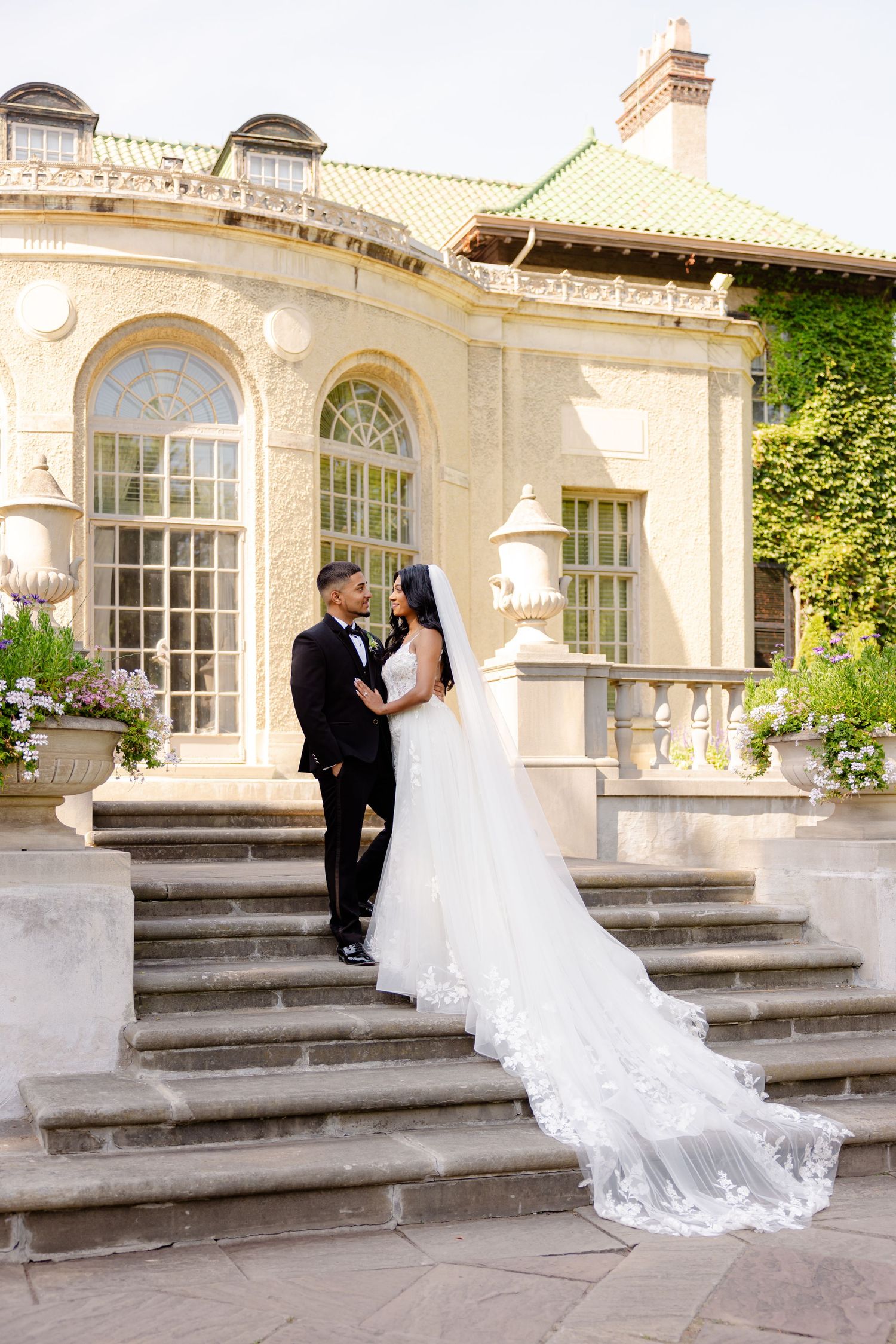 A wedding couple poses on stone steps with a flowing veil against a cream-colored historic building backdrop.