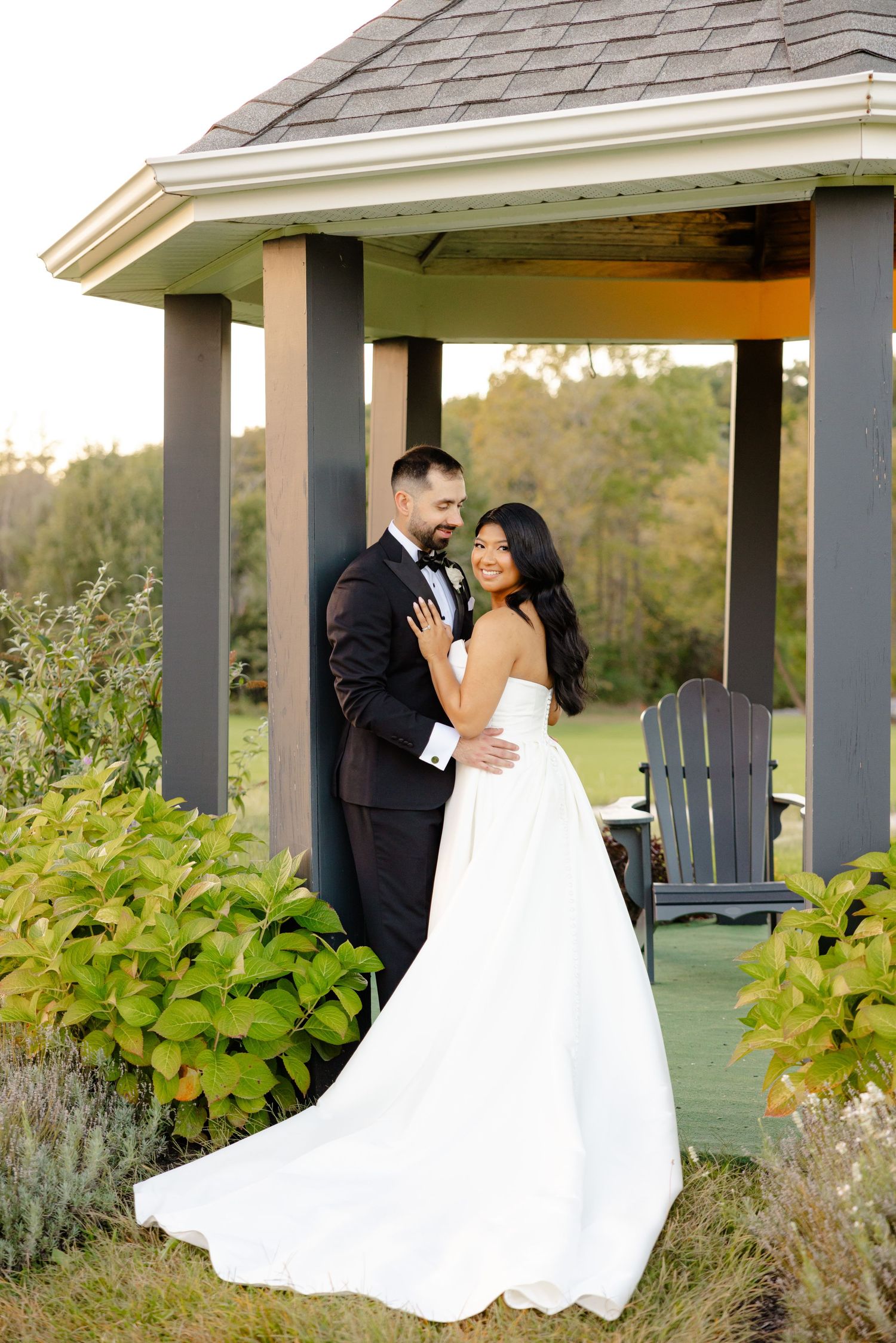A couple in wedding attire shares a tender moment in a garden gazebo at sunset.