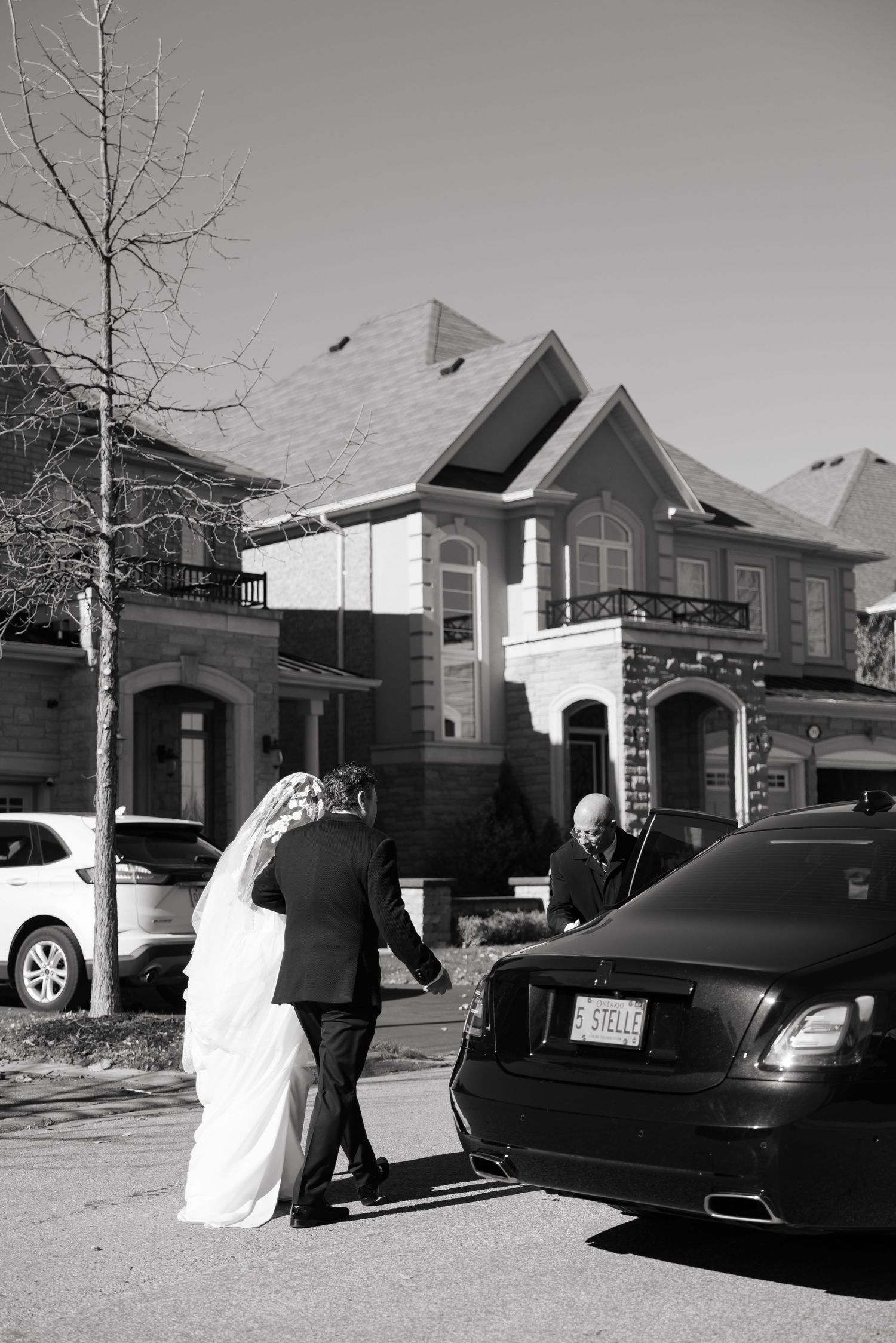Black and white photo of wedding couple entering car outside luxury homes.