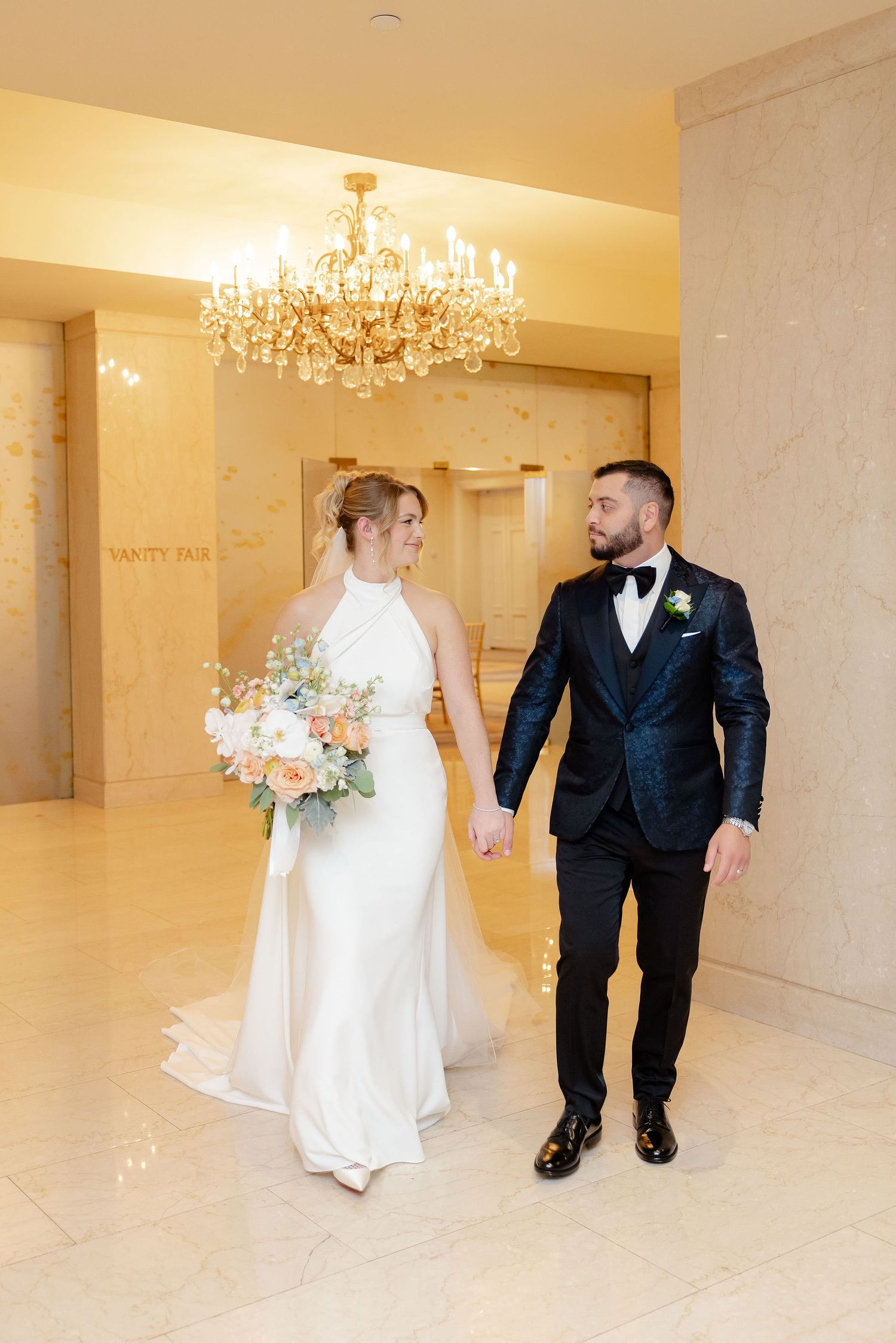 Bride and groom stroll through an elegant venue with crystal chandeliers and cream-colored marble walls.