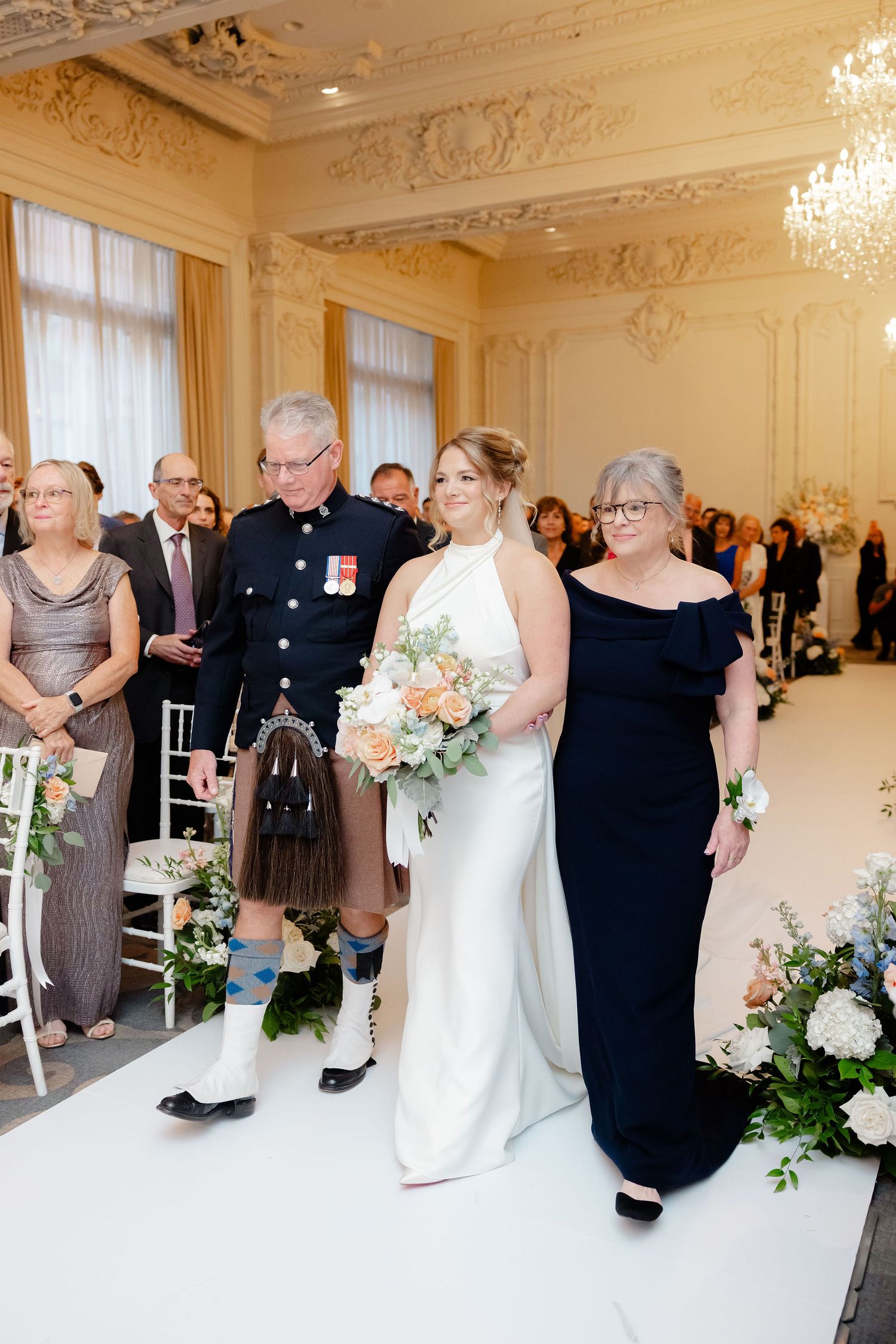 Wedding guests gather in an elegant ballroom with ornate moldings and a chandelier during a formal celebration.