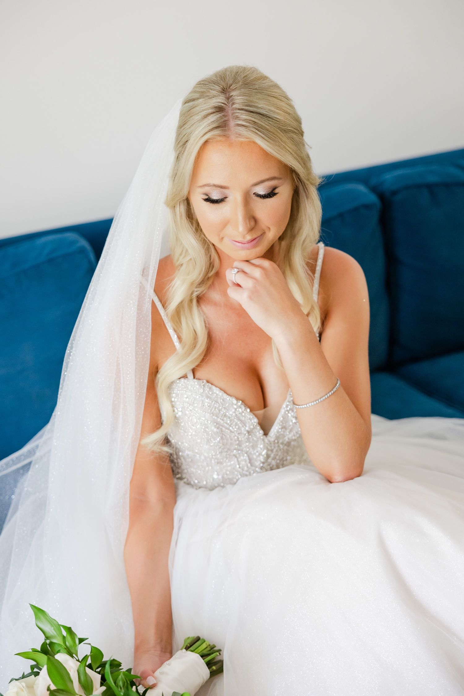 Bride in embellished sweetheart gown poses on blue sofa with flowing veil.
