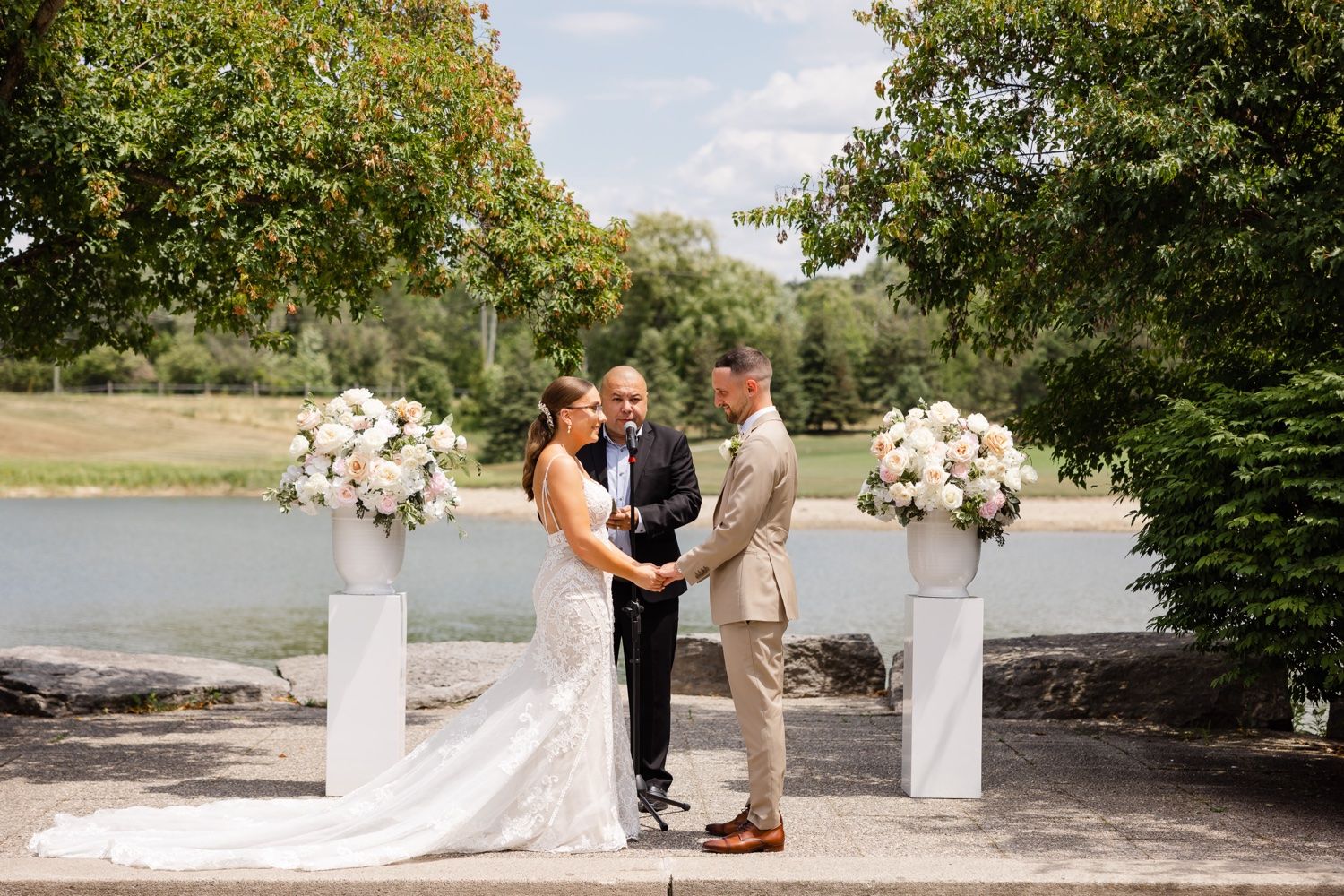 Outdoor wedding ceremony by a lake with white floral arrangements and natural greenery.
