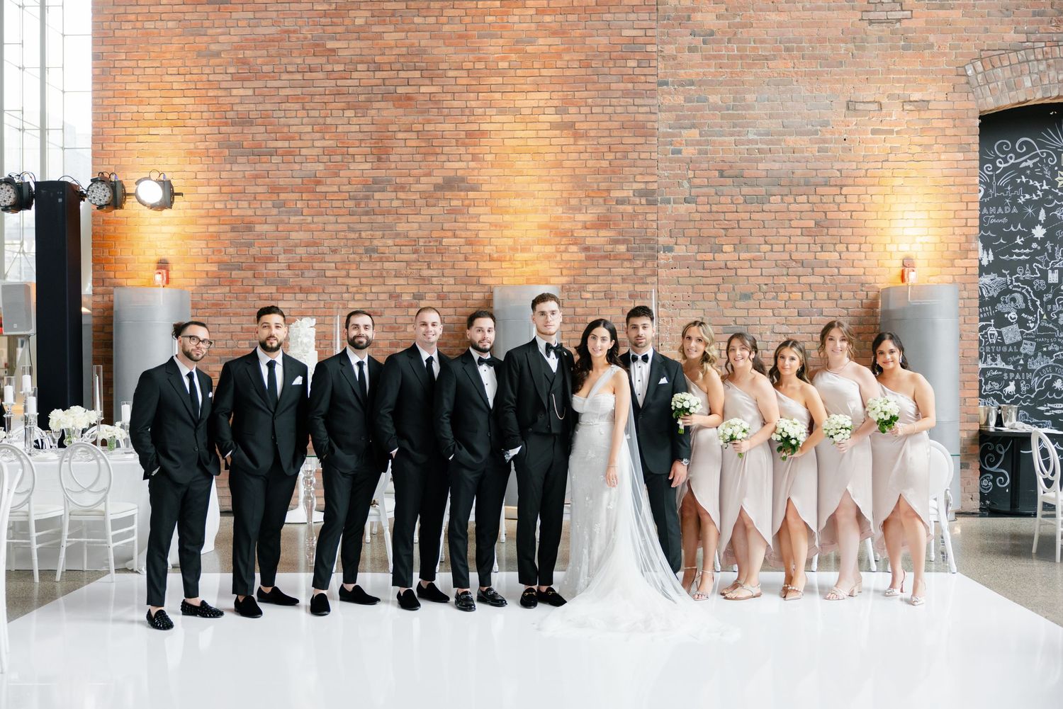 Full wedding party in formal attire poses against brick wall at modern reception venue.
