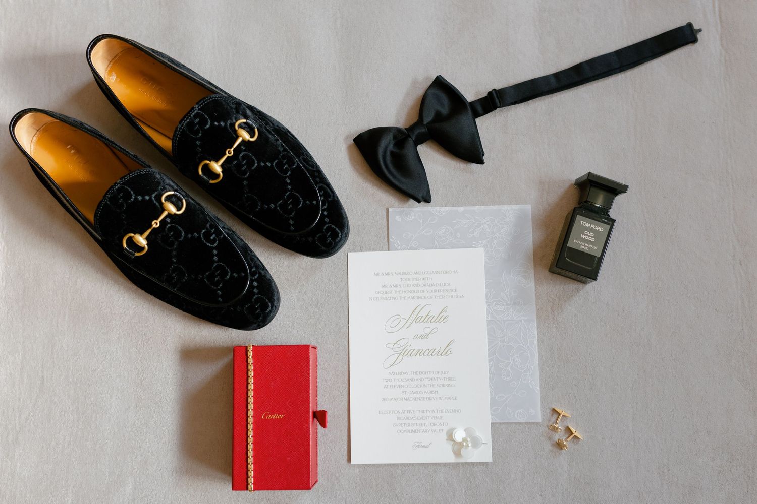 Wedding details flatlay with black leather loafers, invitation, and accessories on light fabric.
