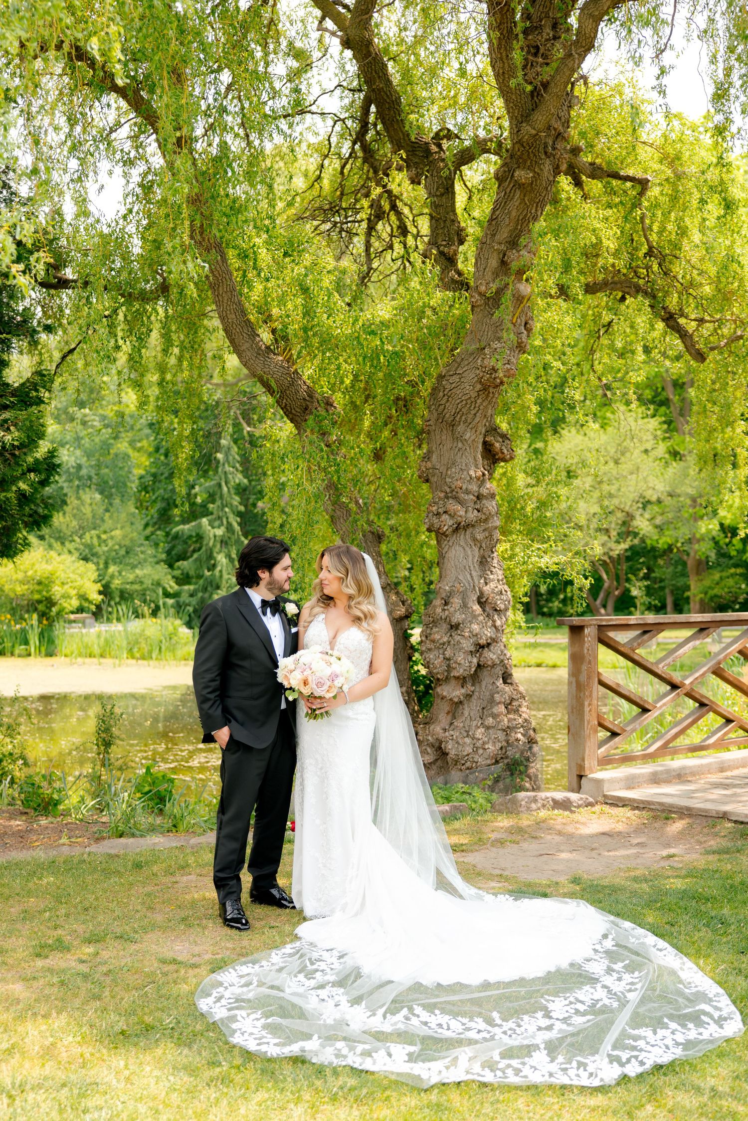 Newlyweds stand together under willow tree with ornate lace veil flowing across garden path.