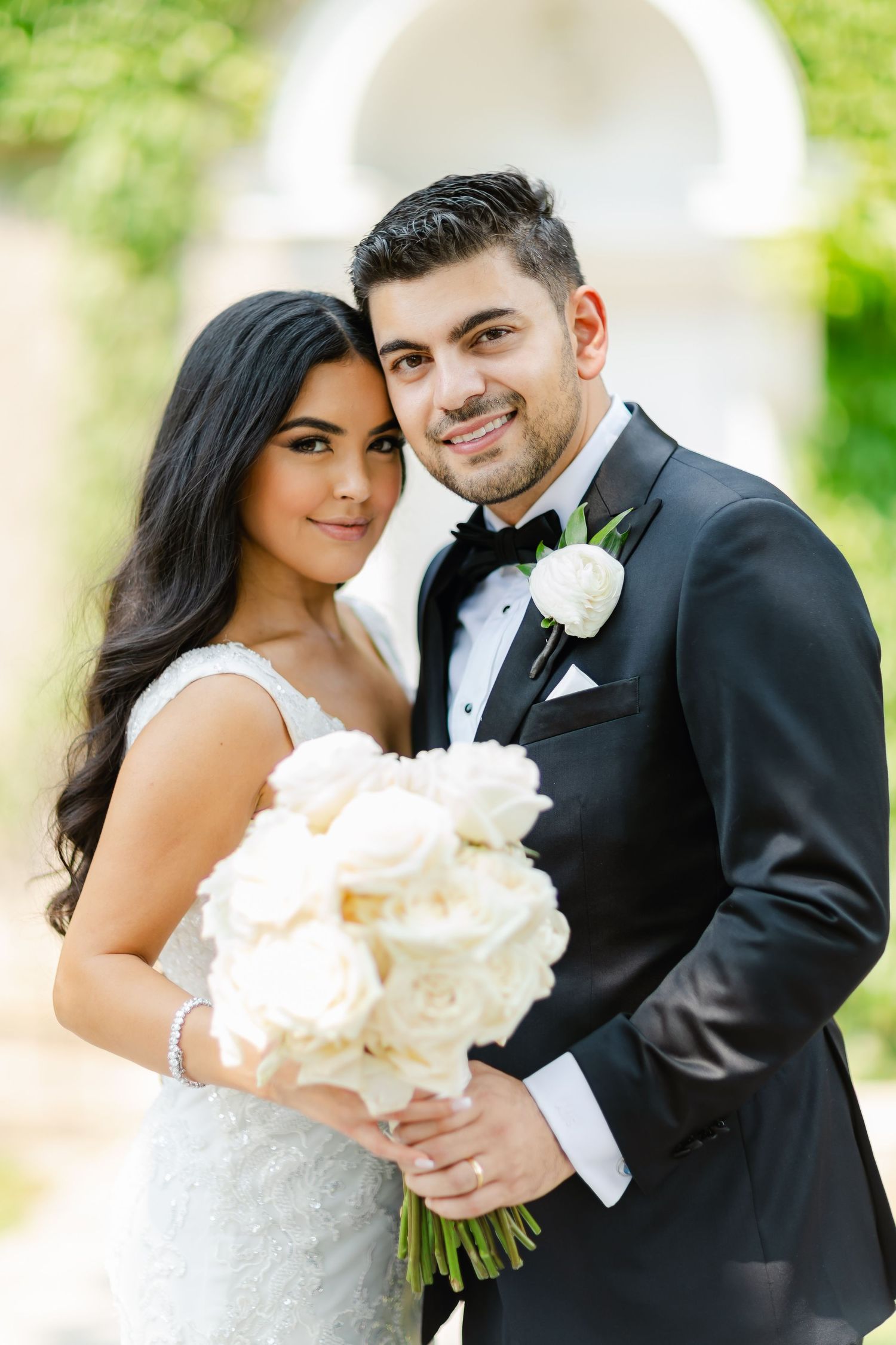 Newlyweds share intimate moment while holding white rose bouquet against garden archway.