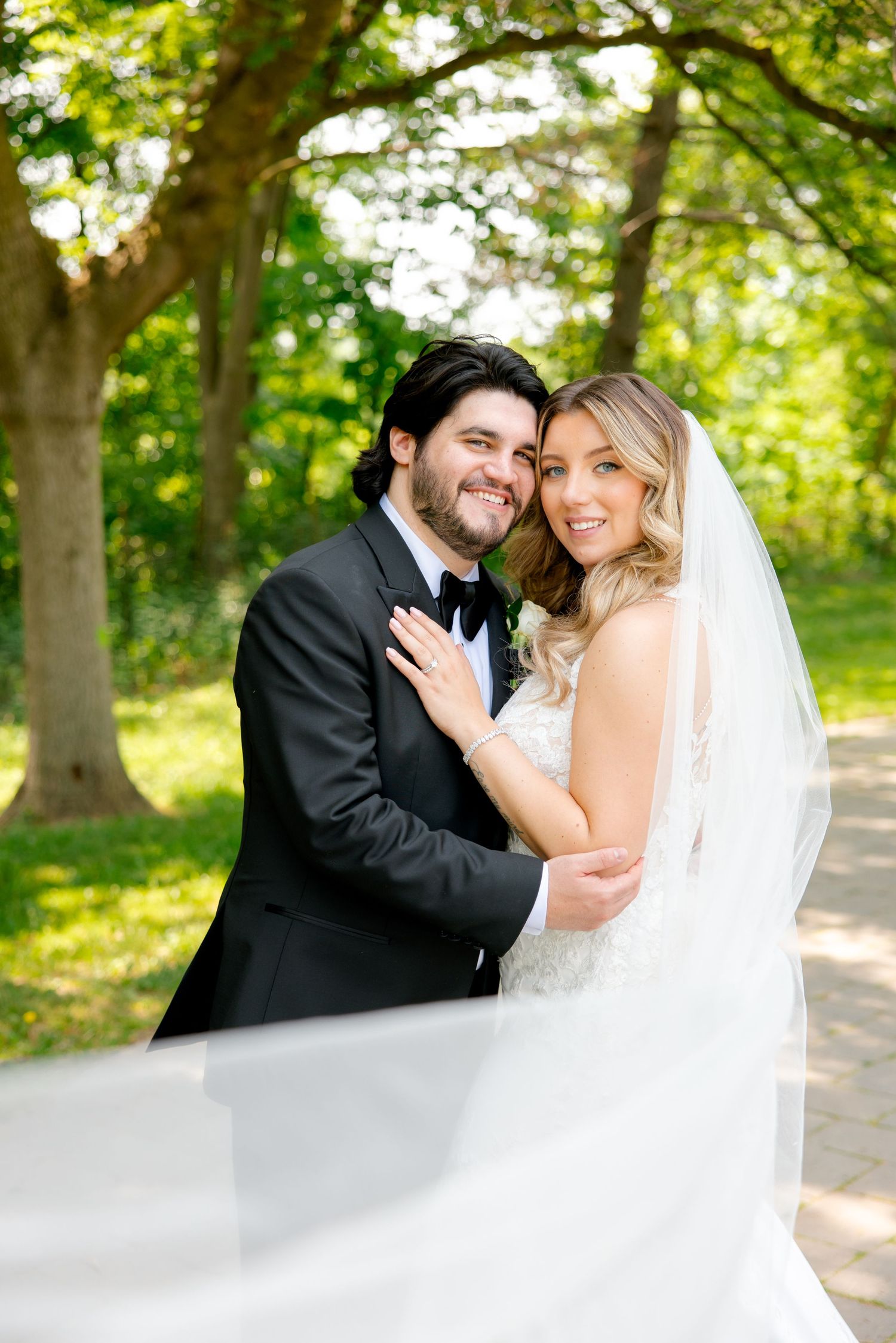 Sunlit outdoor wedding portrait under lush green trees.
