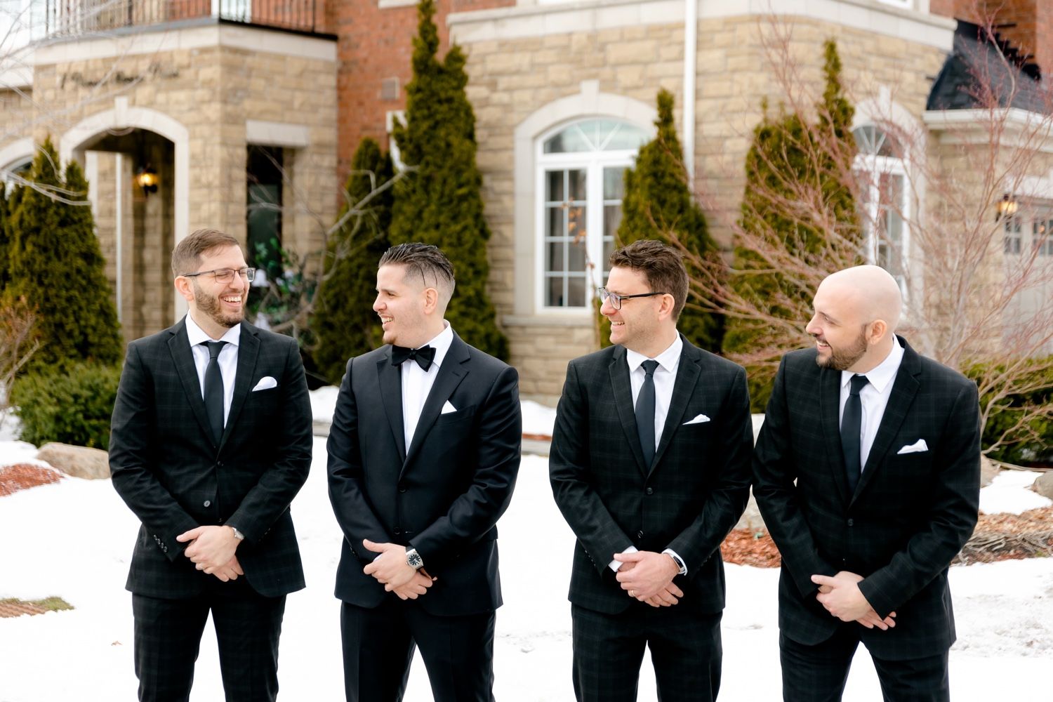 Groomsmen share laughs while standing in front of elegant stone mansion.