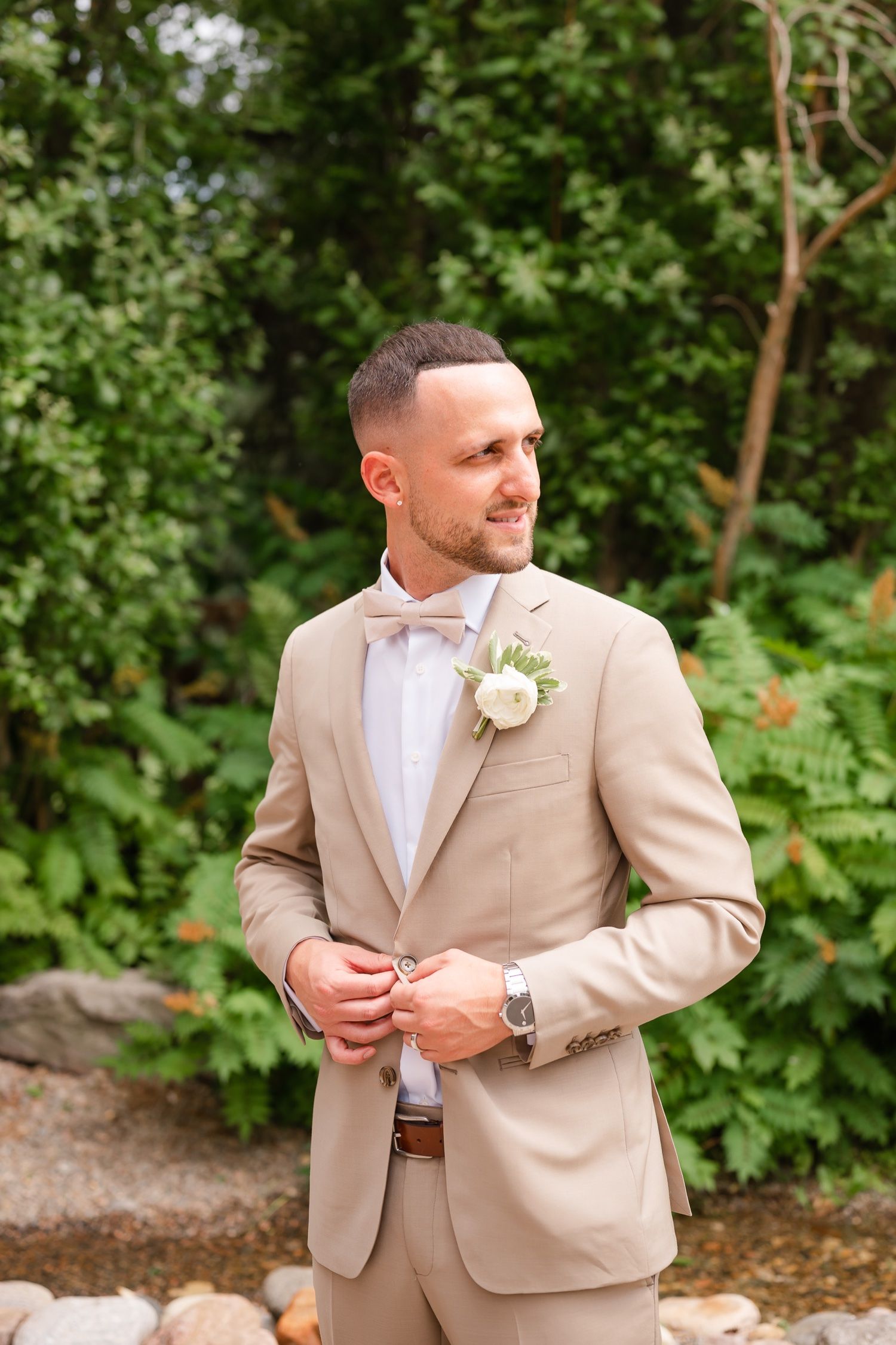 Sharp dressed groom in beige suit with boutonniere stands outdoors against lush greenery background.