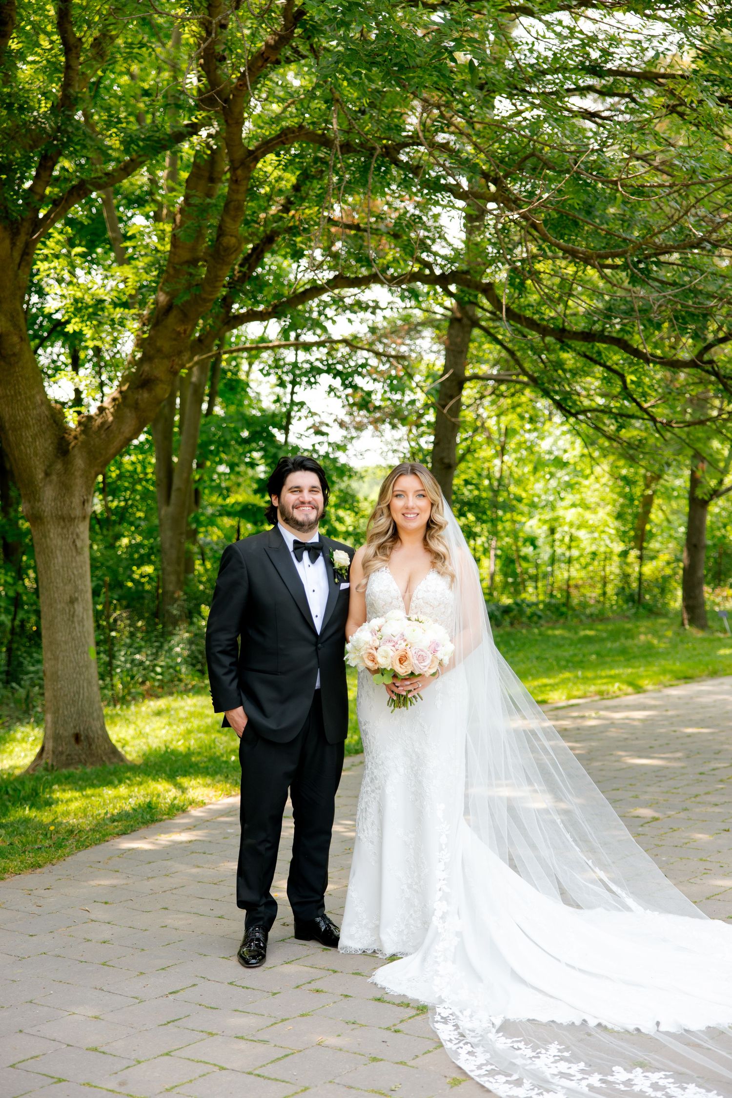 A bride and groom pose on a tree-lined path, with her long lace veil trailing behind.