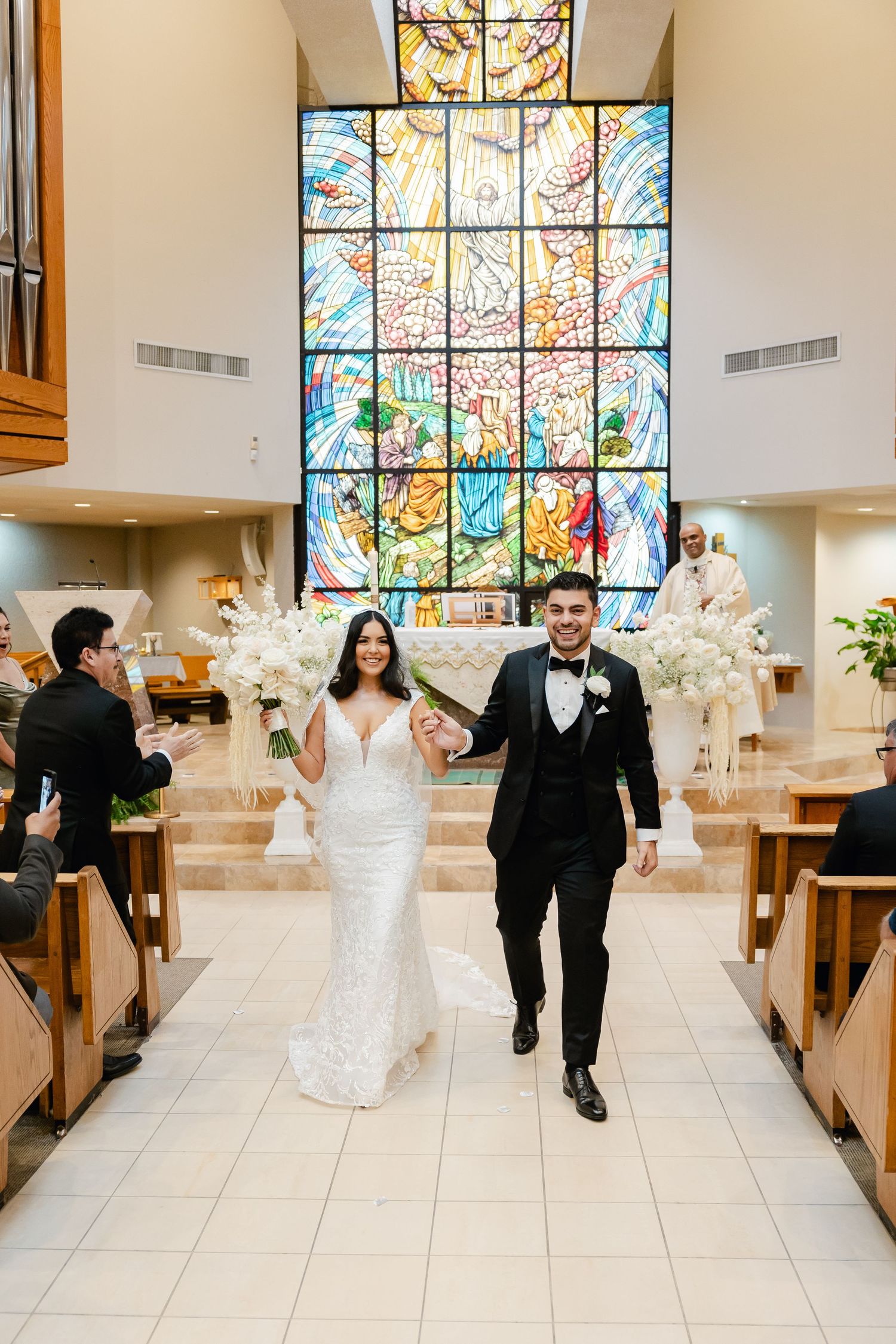 Newlyweds walking down church aisle with colorful stained glass window and white floral decorations.