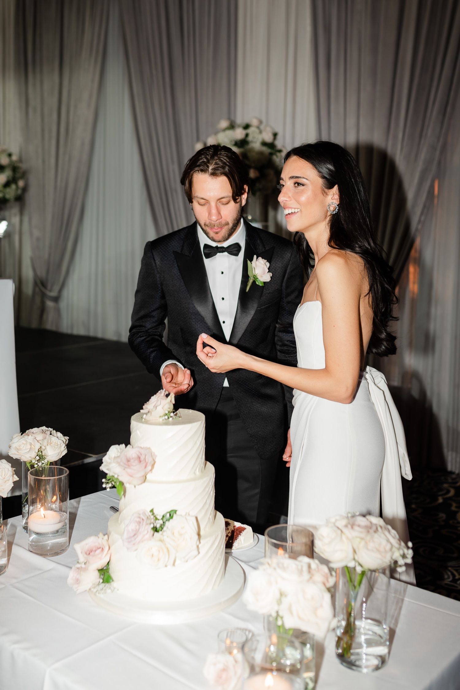 Couple shares tender moment while cutting their three-tiered white wedding cake decorated with pink flowers.