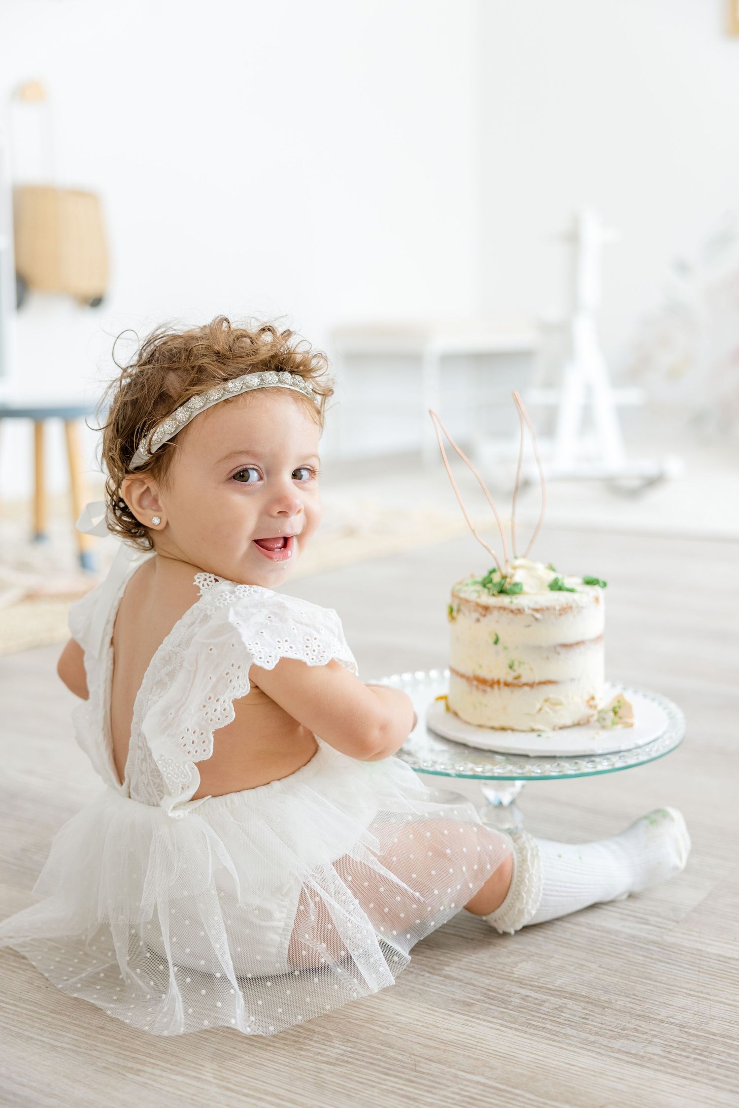 Small child wearing white lace dress and headband sits near decorated cake on wooden floor.
