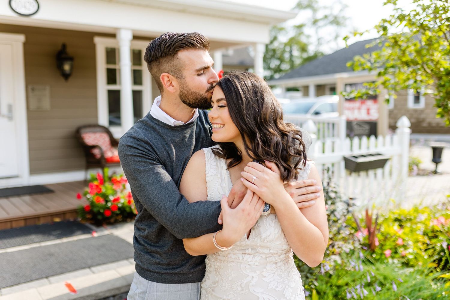 A couple embraces tenderly on their front porch with colorful flowers in the background.