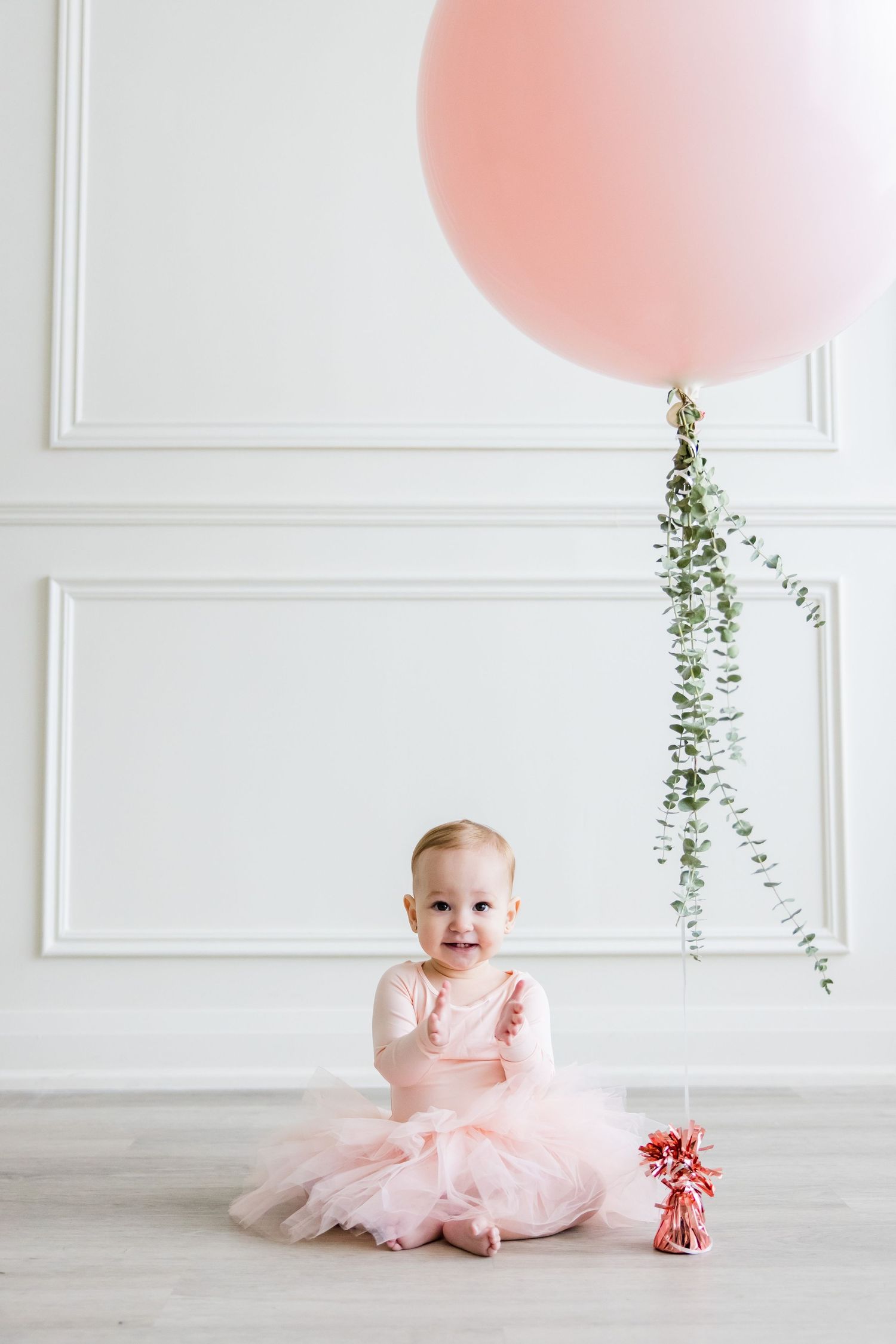 Toddler wearing pink tulle dress sits on floor with large pink balloon and greenery decor.