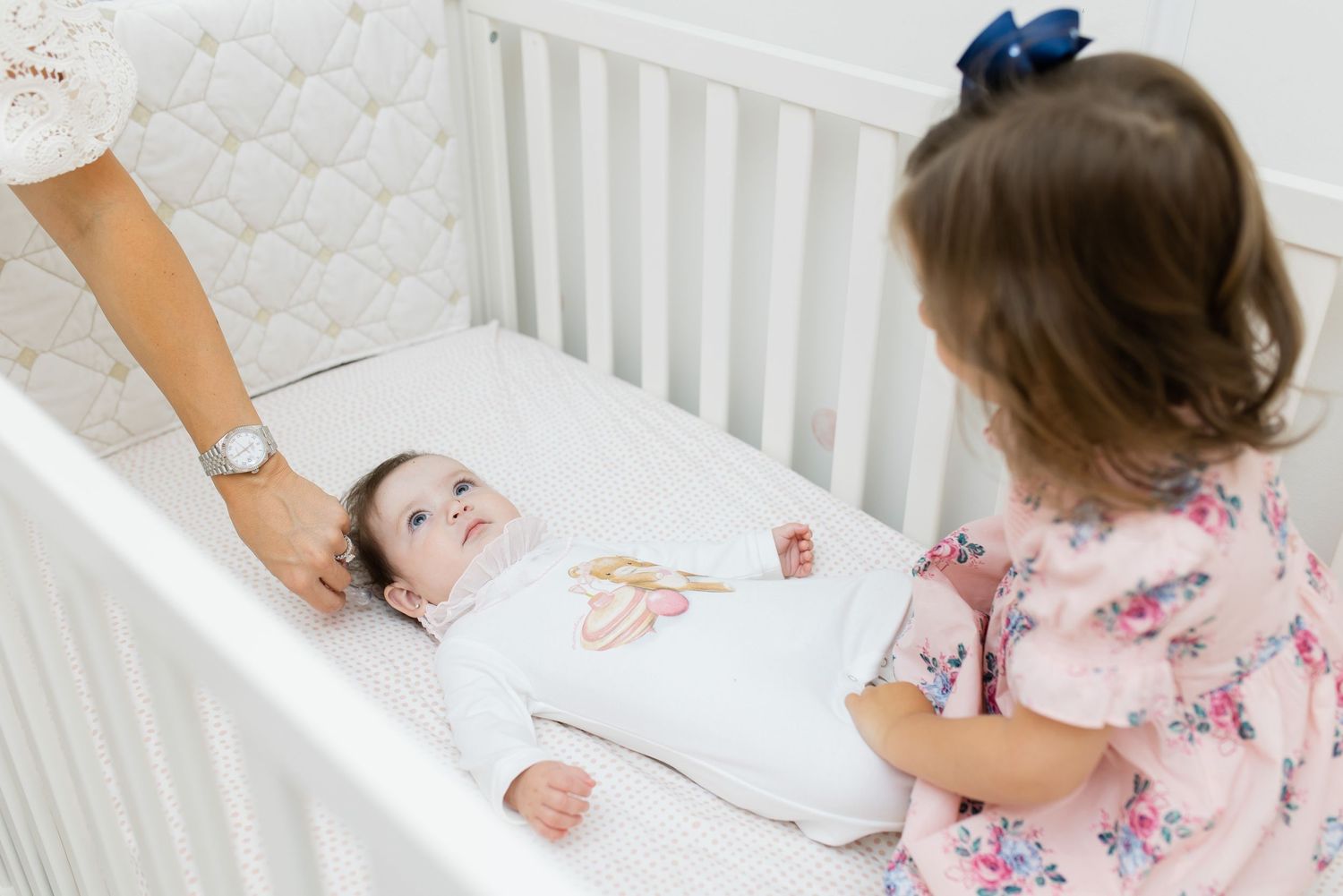 A baby in white lies in a crib while being watched by someone in a pink floral dress.