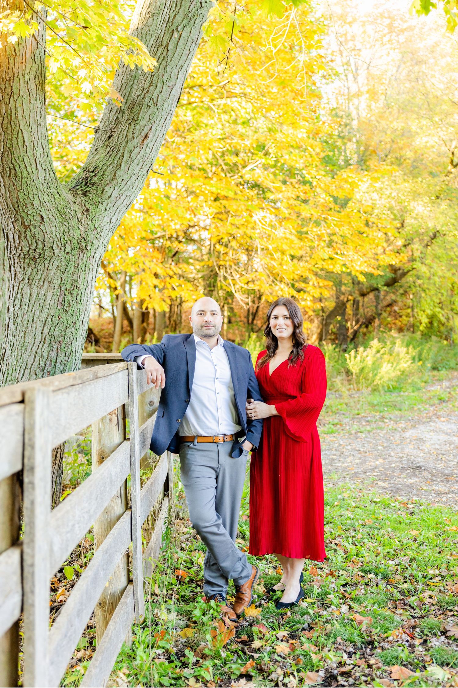 Elegant couple in formal wear standing by wooden fence with autumn trees overhead.