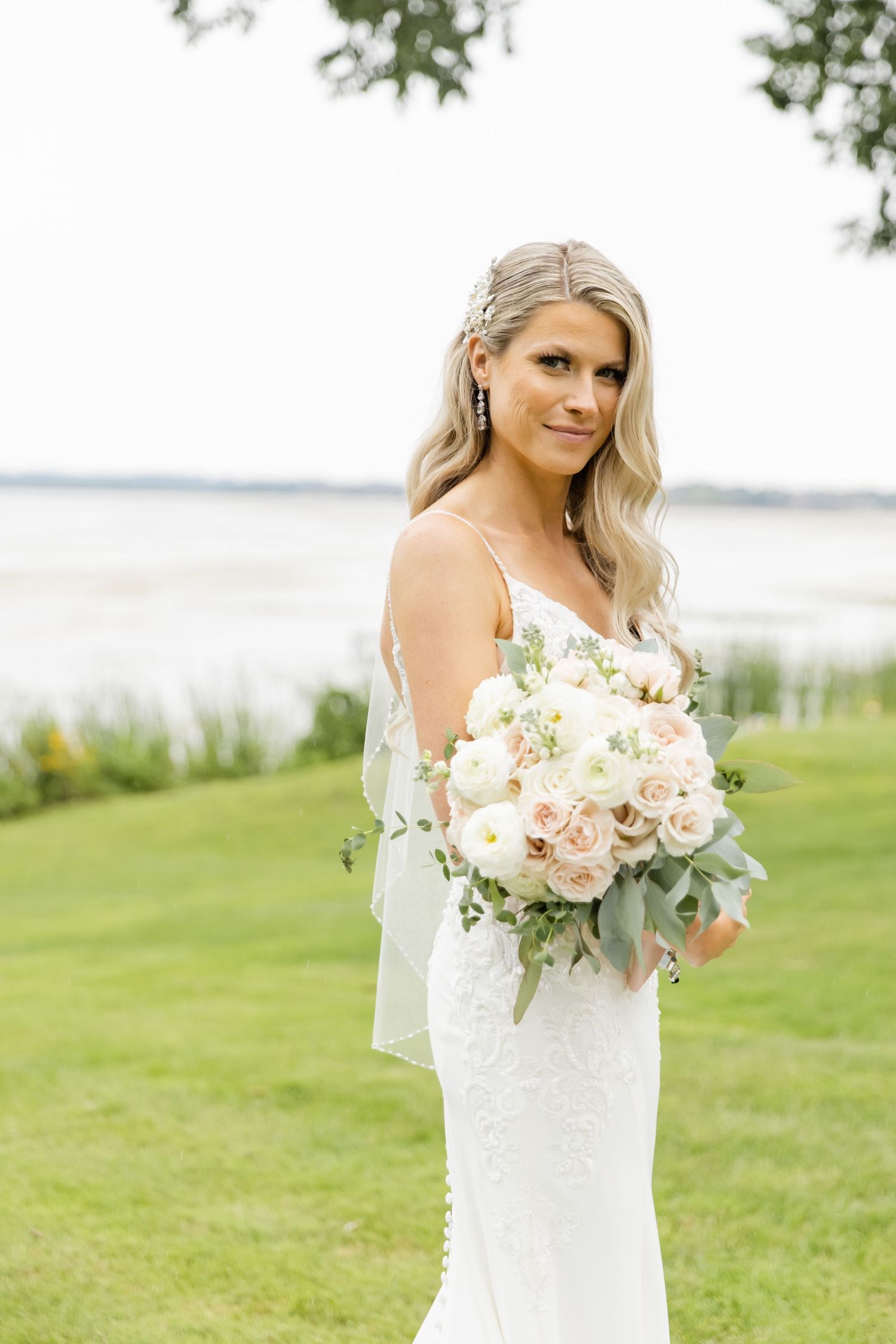 Elegant bride in fitted lace gown holds blush and white rose bouquet by waterfront.
