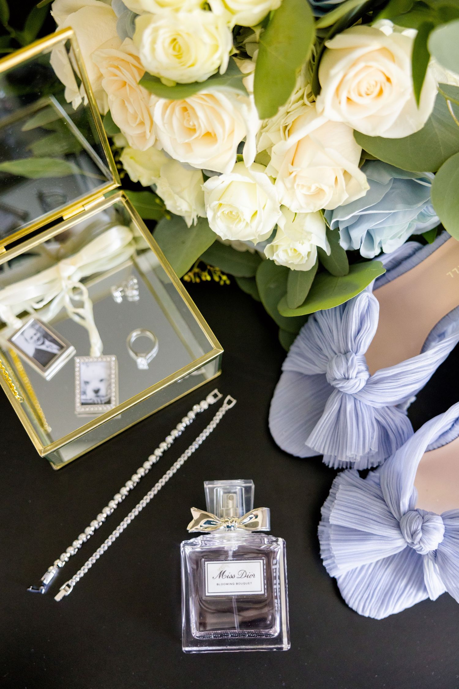 Wedding accessories with white roses, crystal necklace, perfume bottle and blue bow shoes on black surface.